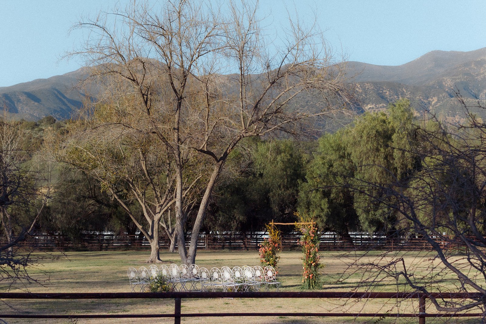 An outdoor wedding setup with white chairs arranged in a row, decorated arch with flowers, and trees with mountains in the background.