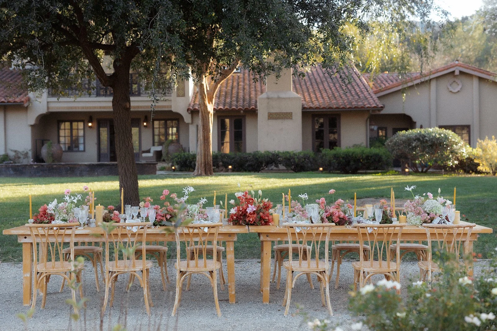 A long wooden banquet table set outdoors with pink and white floral centerpieces, candles, and glassware, arranged in front of a large house with a red tile roof and lush landscaping, under the shade of tall trees.