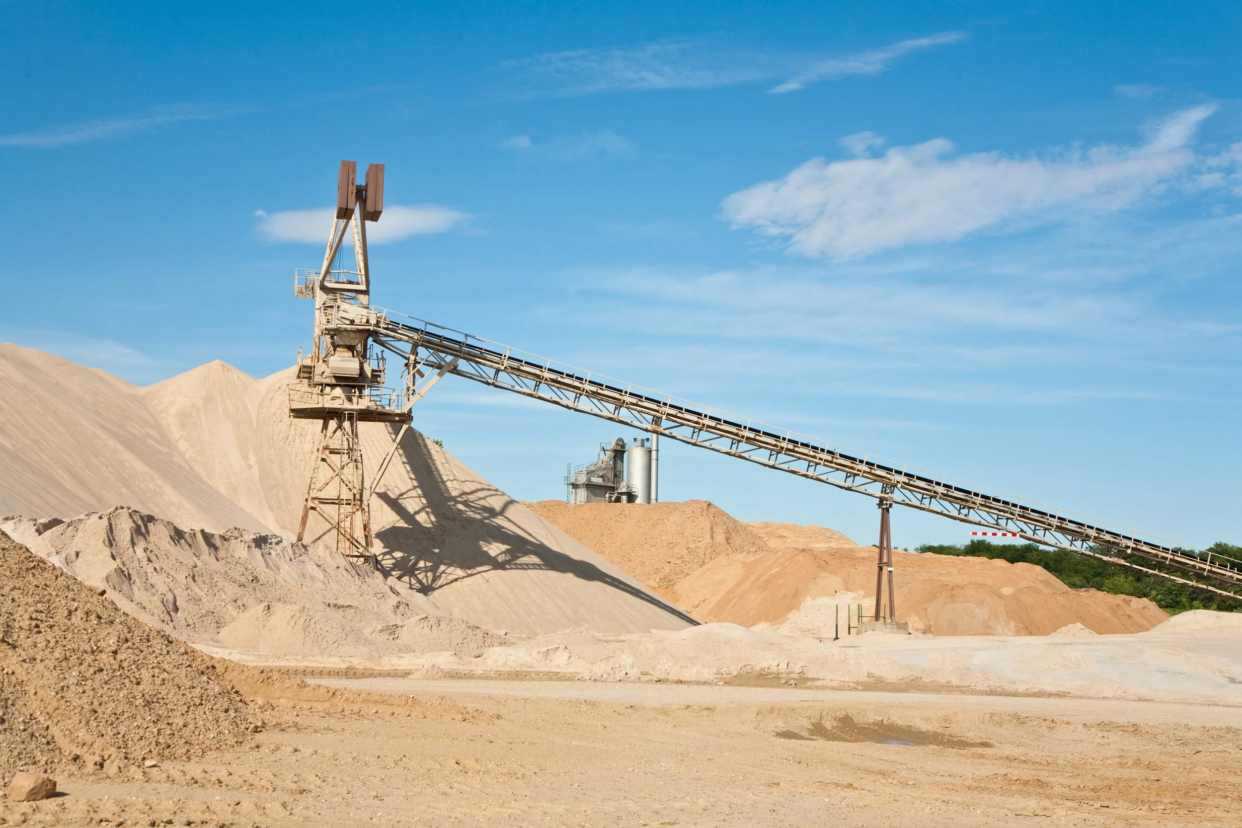 An open-pit sand and gravel mining site with a conveyor belt to transport materials, under a blue sky with some clouds.