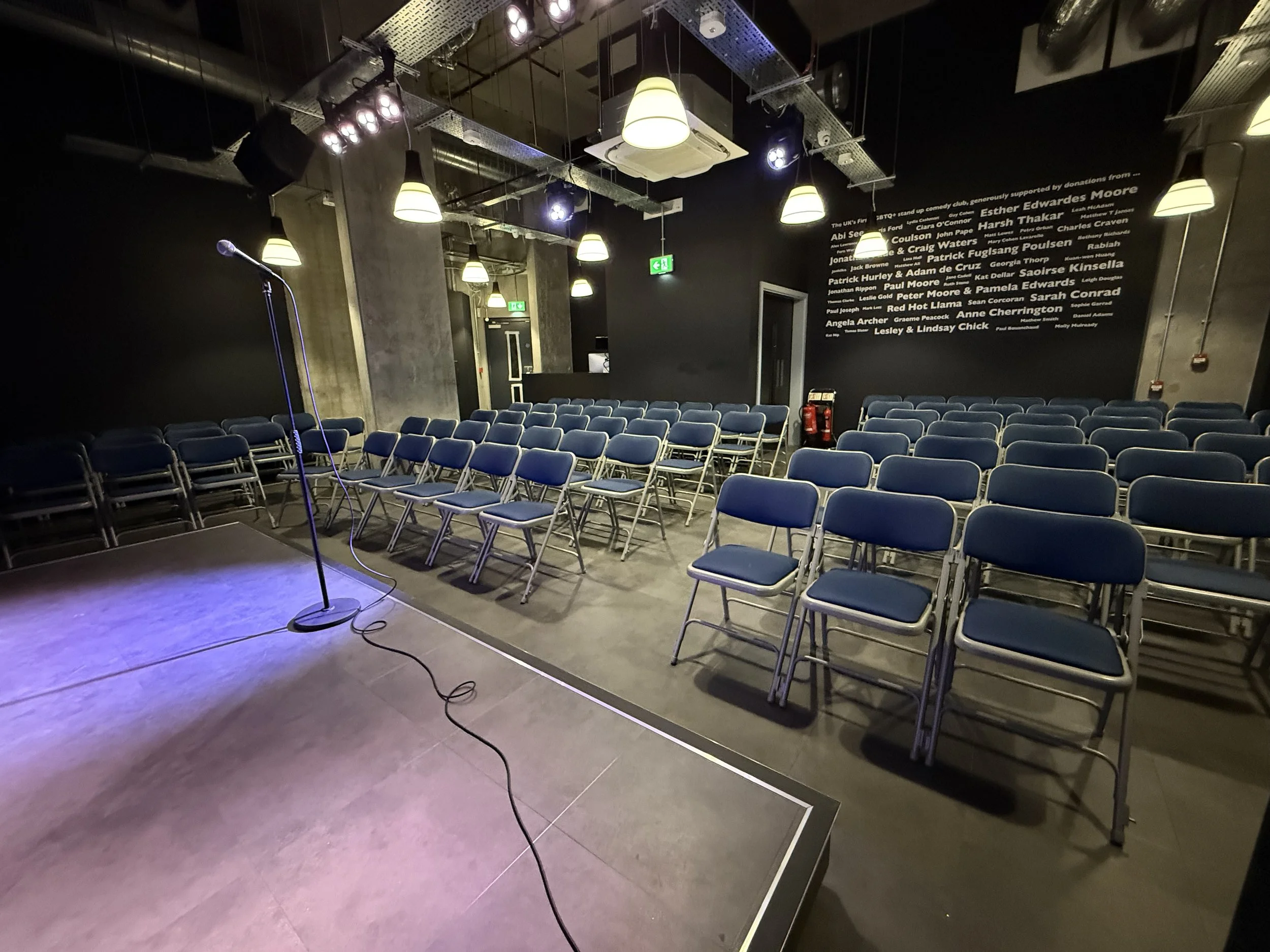 Empty stage with a microphone stand and blue chairs arranged for an event, with a black wall and white text listing names on the background wall.