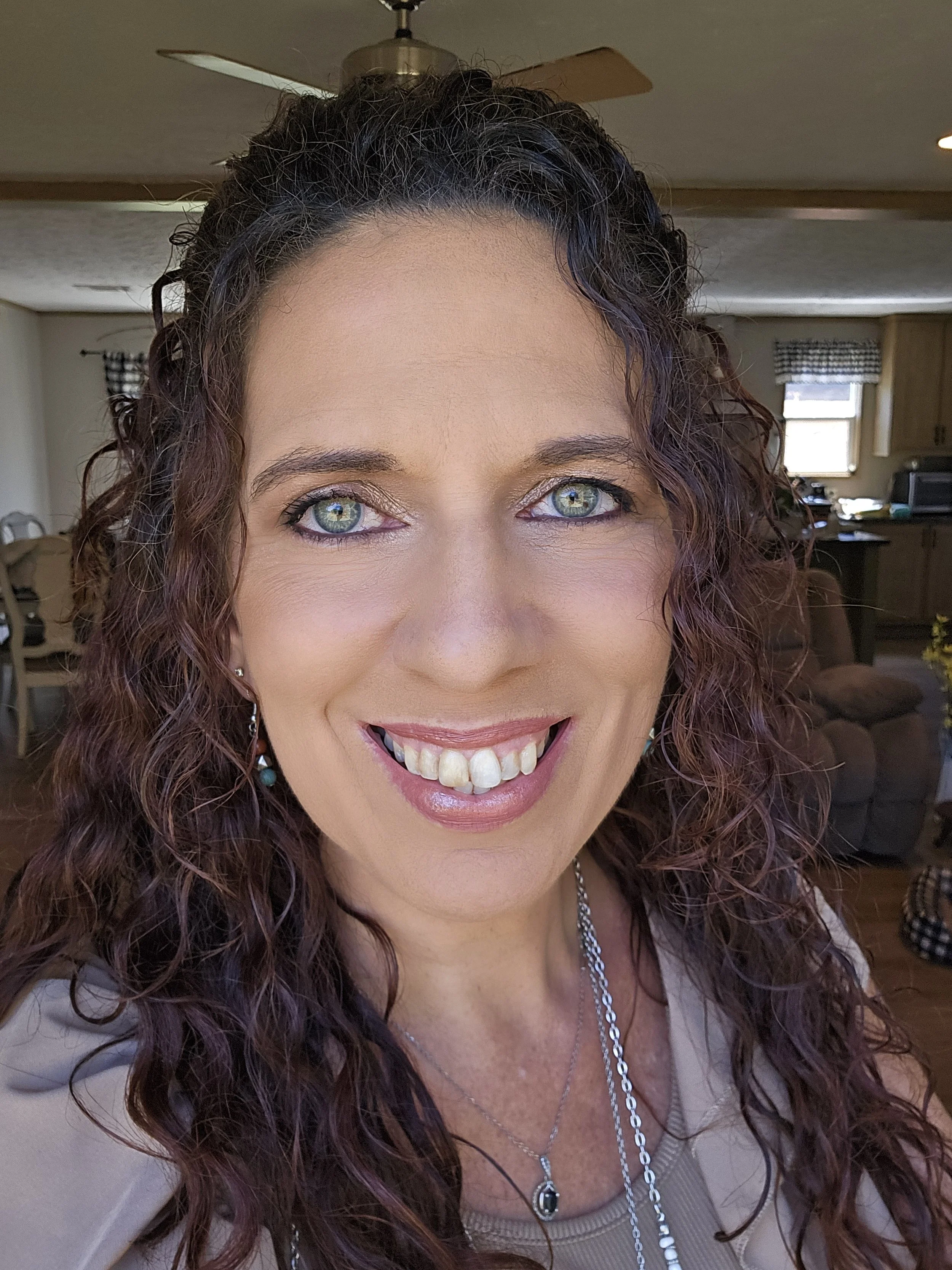 Woman smiling indoors with curly hair, green eyes, and wearing a necklace; background featuring a living room setting with a ceiling fan and furniture.