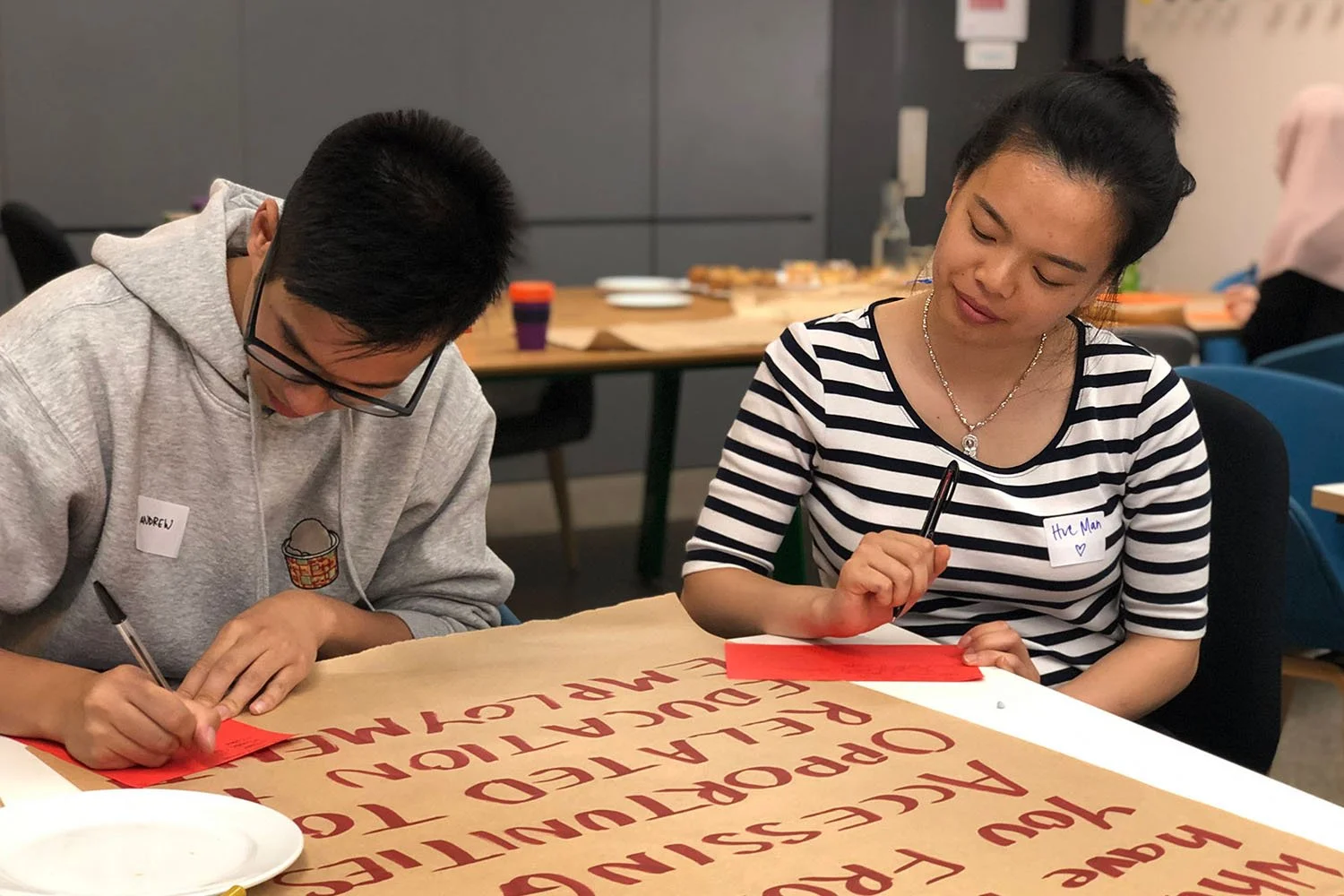 Two young people sit at a table in a co-design workshop. They are both looking down, focused on writing. There is a large piece of butcher's paper on the table with text reading: 'What barriers have prevented you from accessing opportunities related