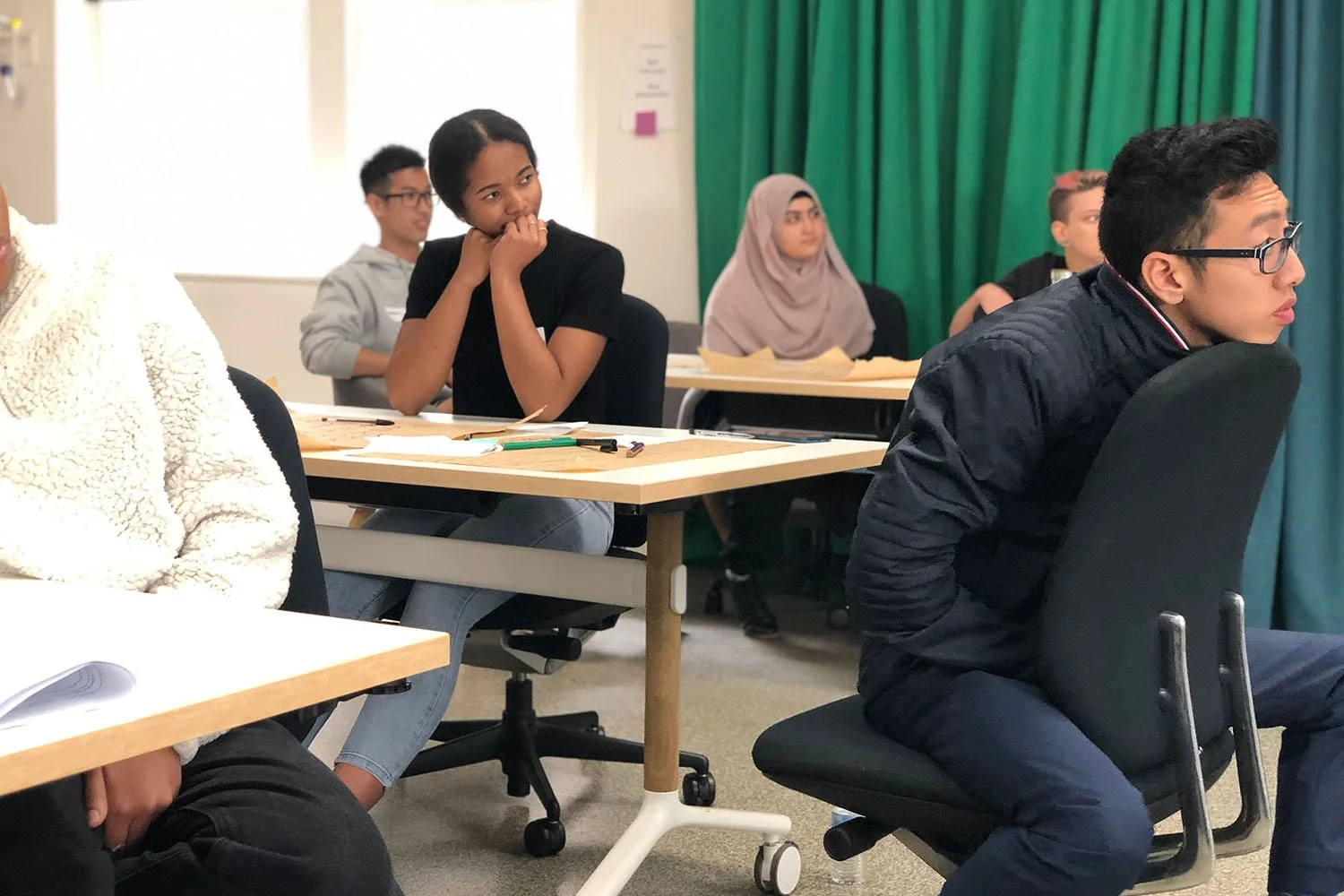 A room full of young people involved in a co-design workshop are sitting at desks and looking off camera, presumably listening to someone speak.