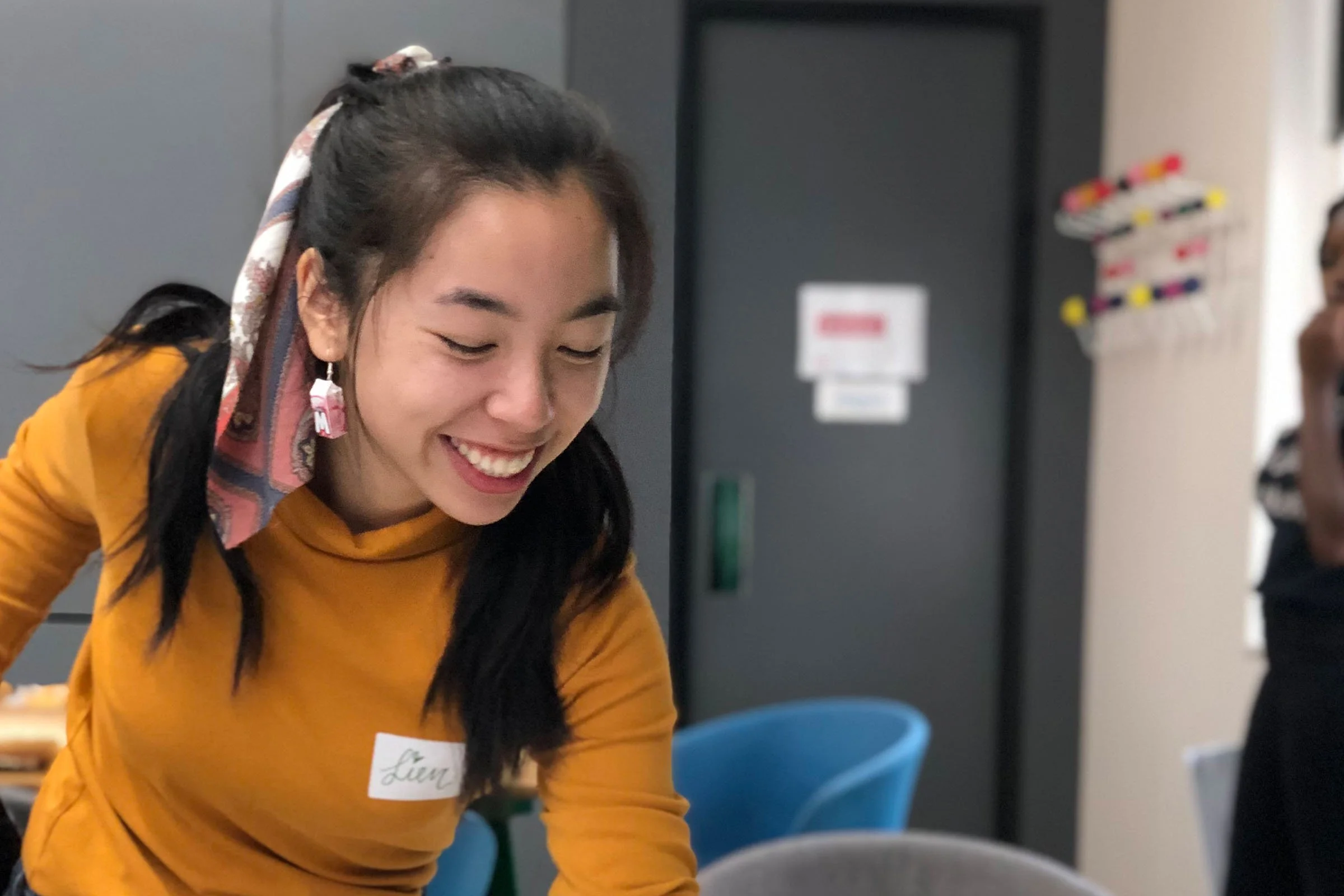A young woman is smiling widely while picking up a yellow piece of paper in a workshop.