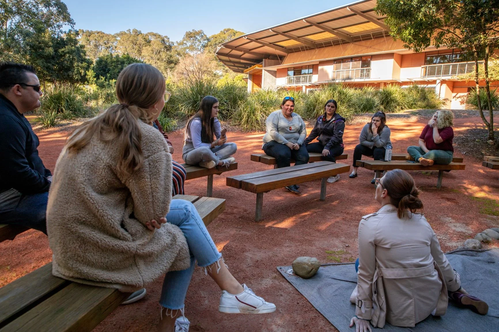 A group of 9 First Nations people sitting outside listening to each other speak at the Wollotuka Institute in Newcastle. There is a building in the distance and the area has red dirt with native grasses and gum trees.