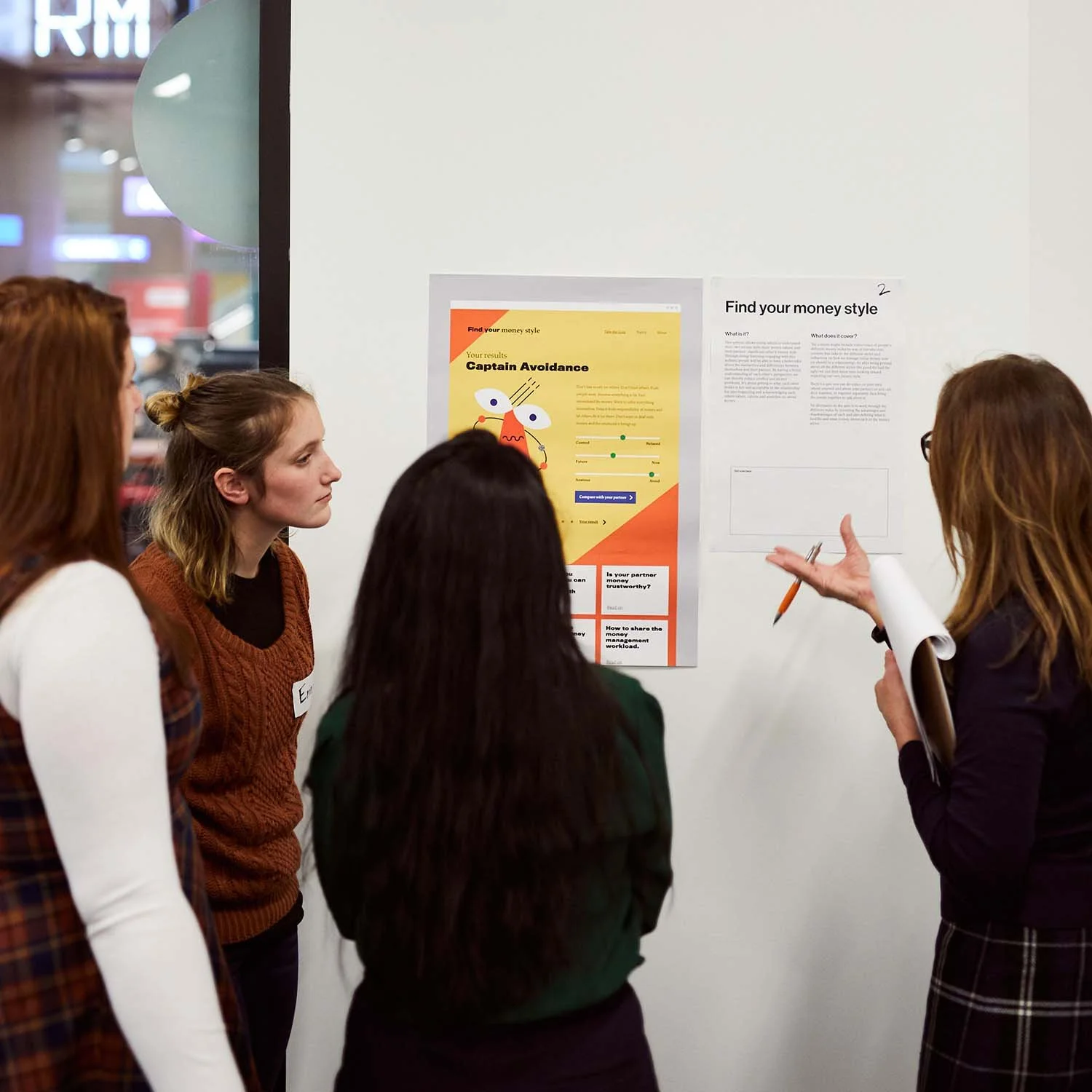A group of four people are gathered around some mock ups for the You, Me & Money website pinned up on the wall. One person is speaking, as shown by gestural hand movements.