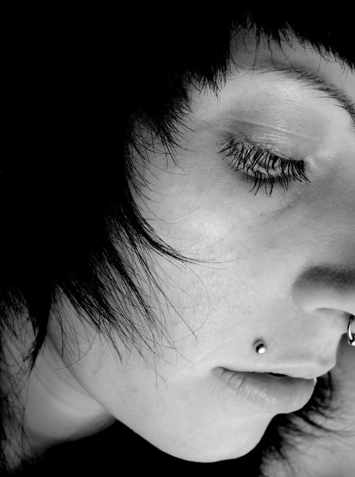 Close-up black and white photo of a young woman with short dark hair, closed eye, long eyelashes, nose piercing, and lip piercing.