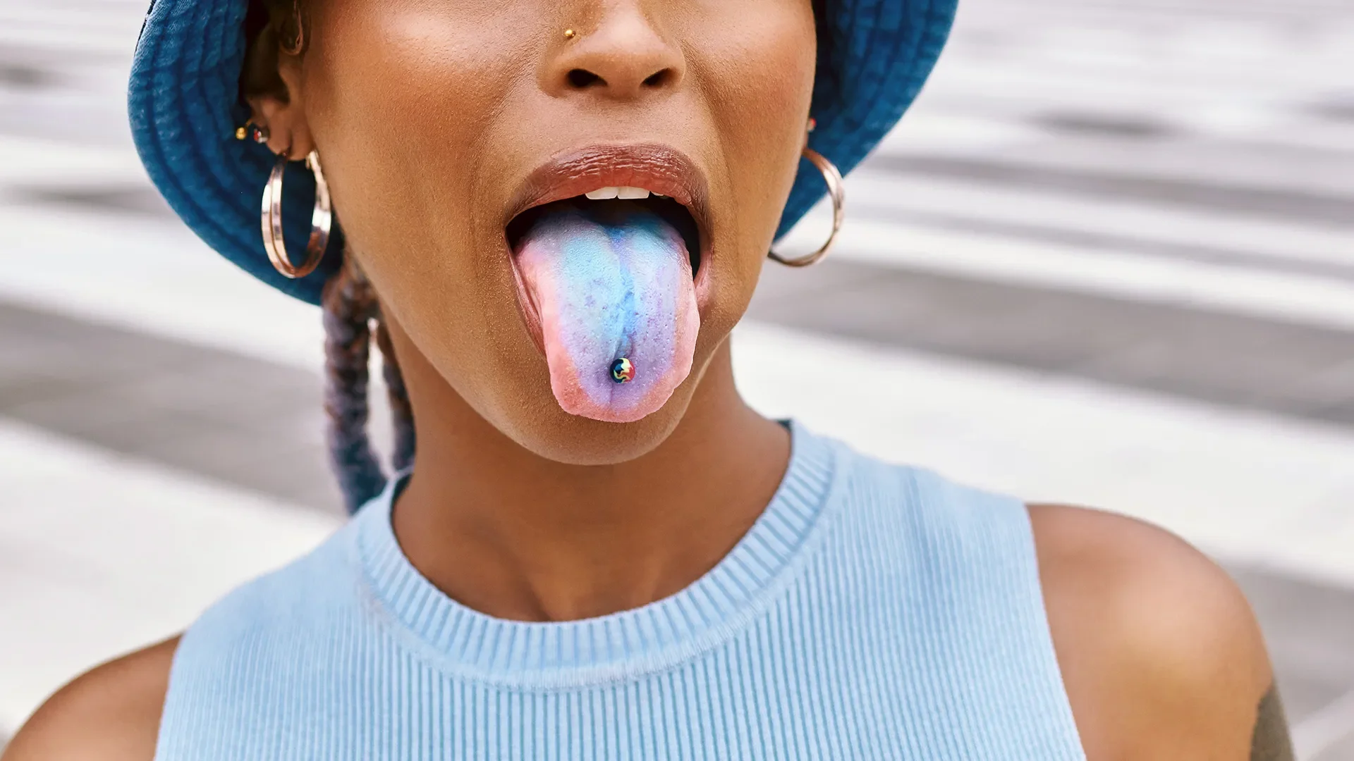 Close-up of a woman with dark skin wearing a blue hat and hoop earrings, sticking out her rainbow-colored tongue with a small rainbow charm piercing, against a blurred background of stairs.