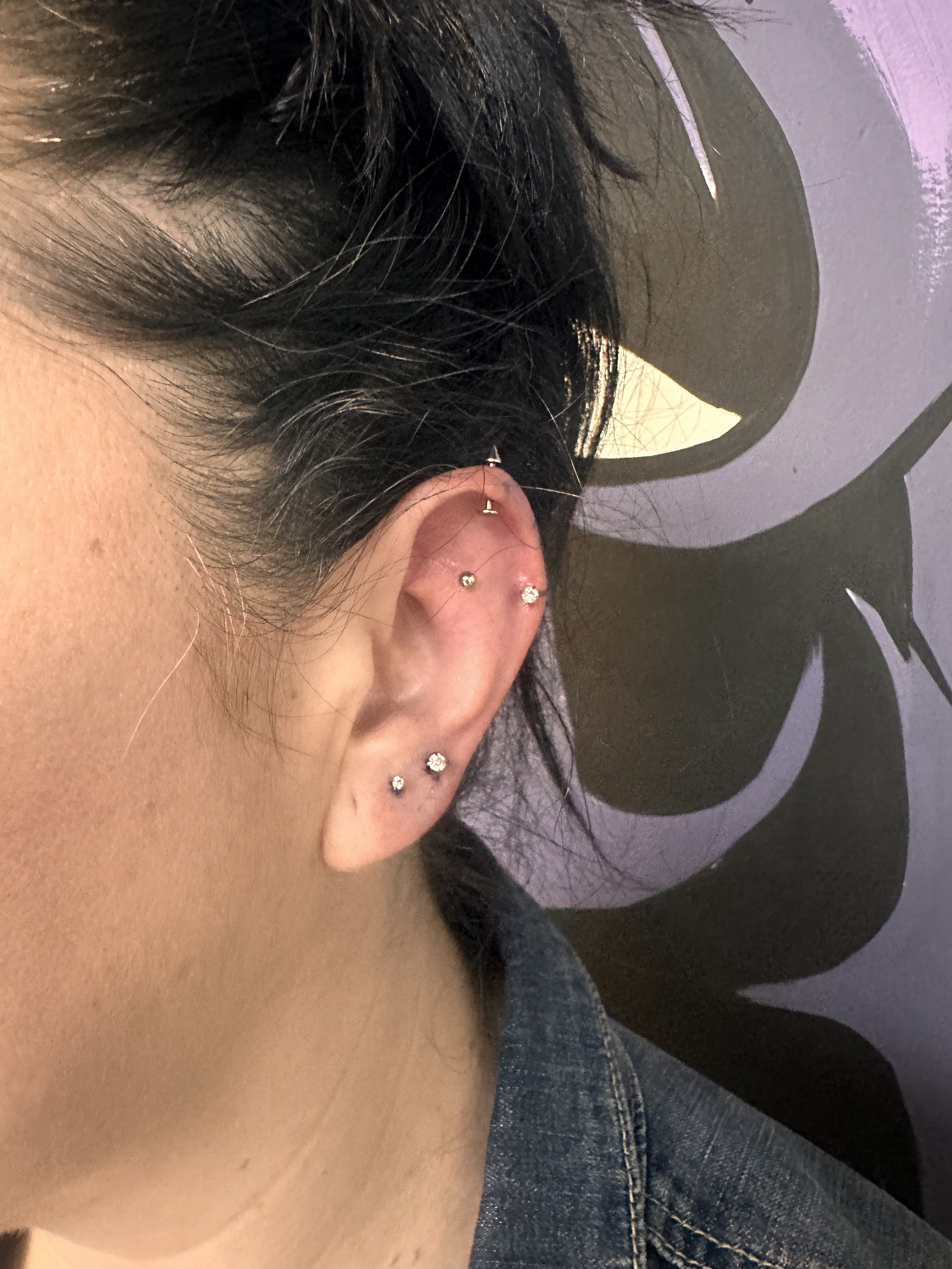 Close-up of a person's ear with multiple piercings, including a helix, tragus, and lobe, wearing small, shiny stud earrings, with short dark hair and a dark background.