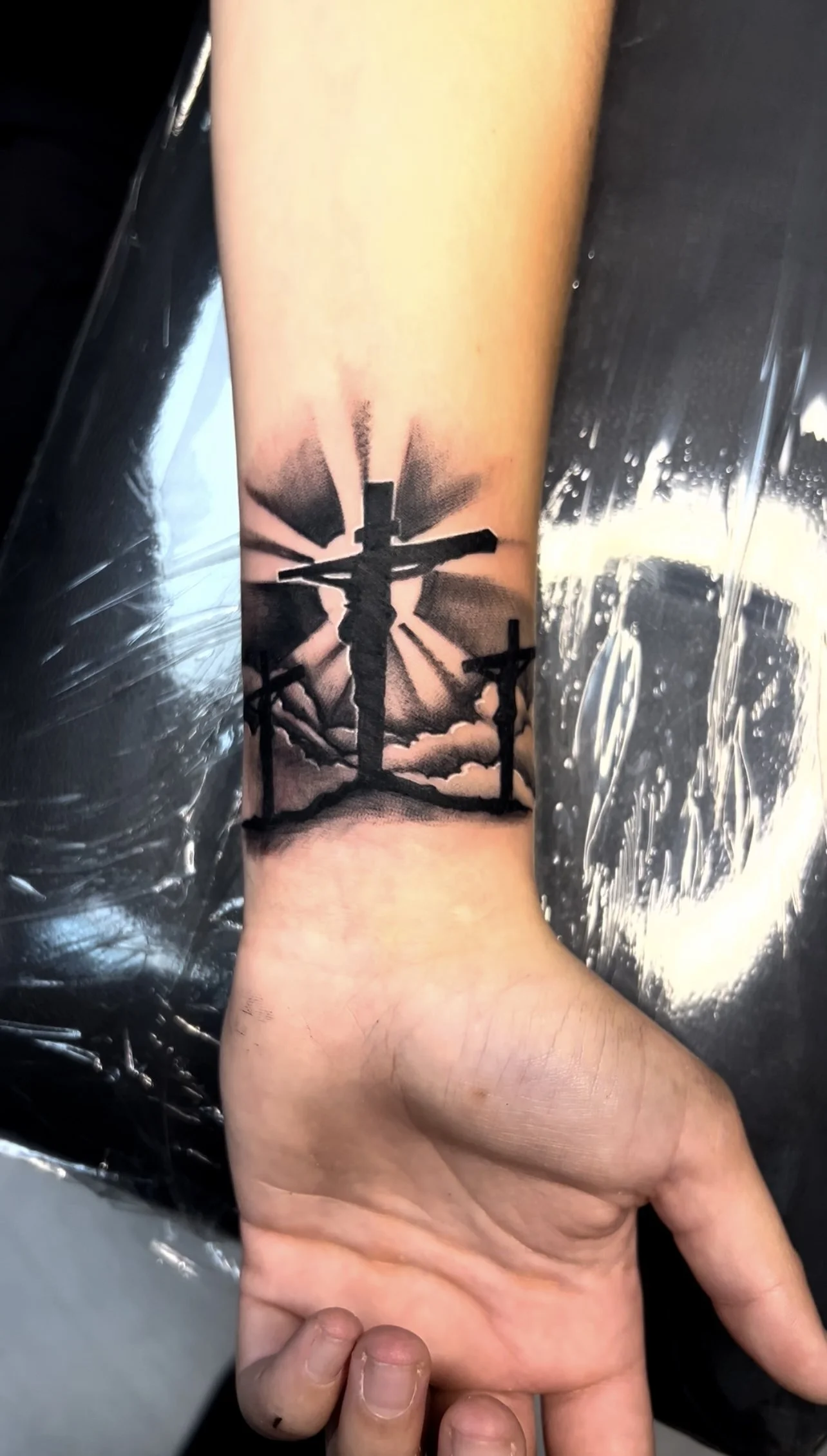Black and gray tattoo of three crosses on person's forearm with a cloudy sky and sun rays in the background.