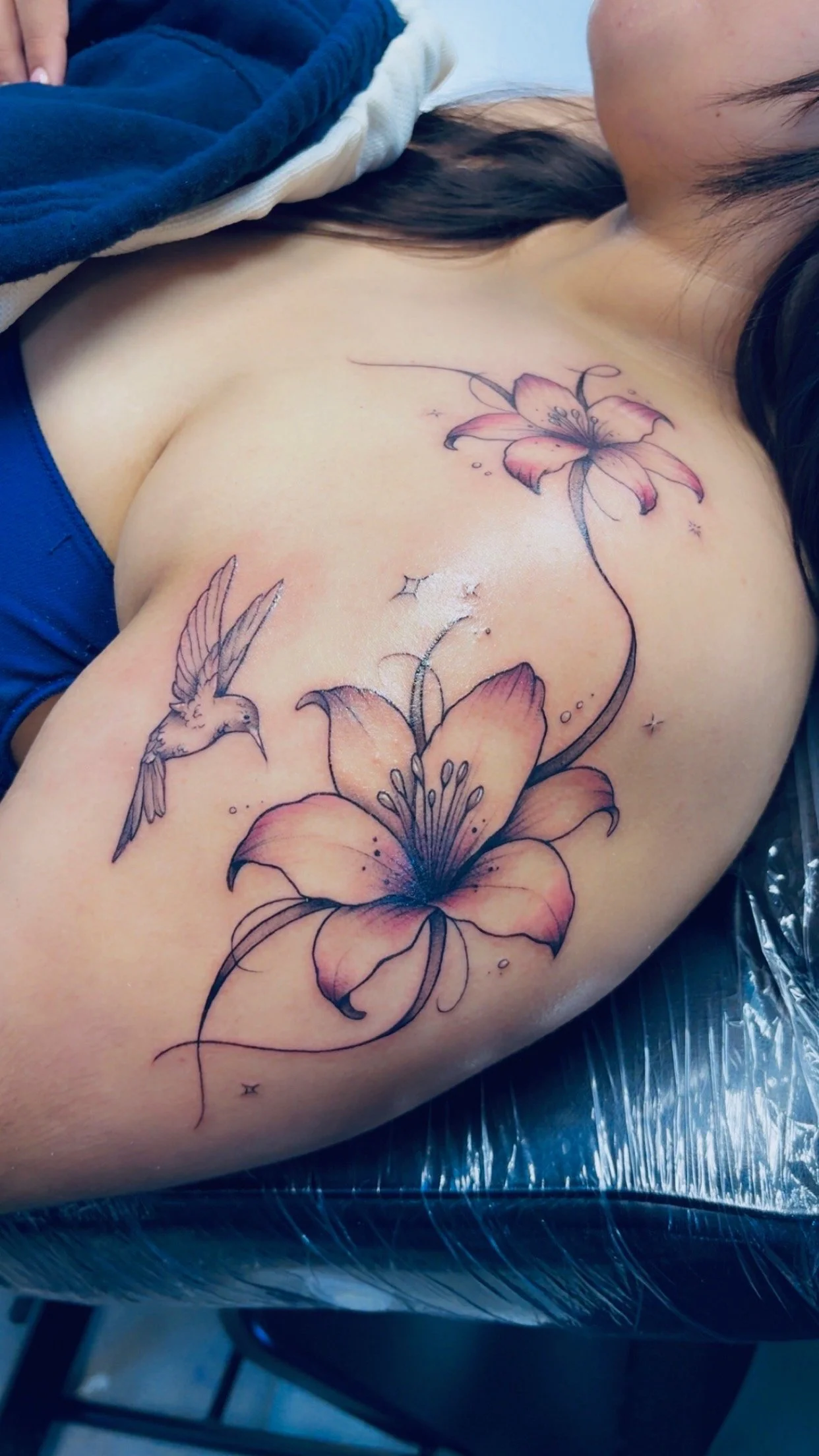 Close-up of a woman's shoulder with a colorful floral tattoo featuring lilies, a hummingbird, and small decorative stars.