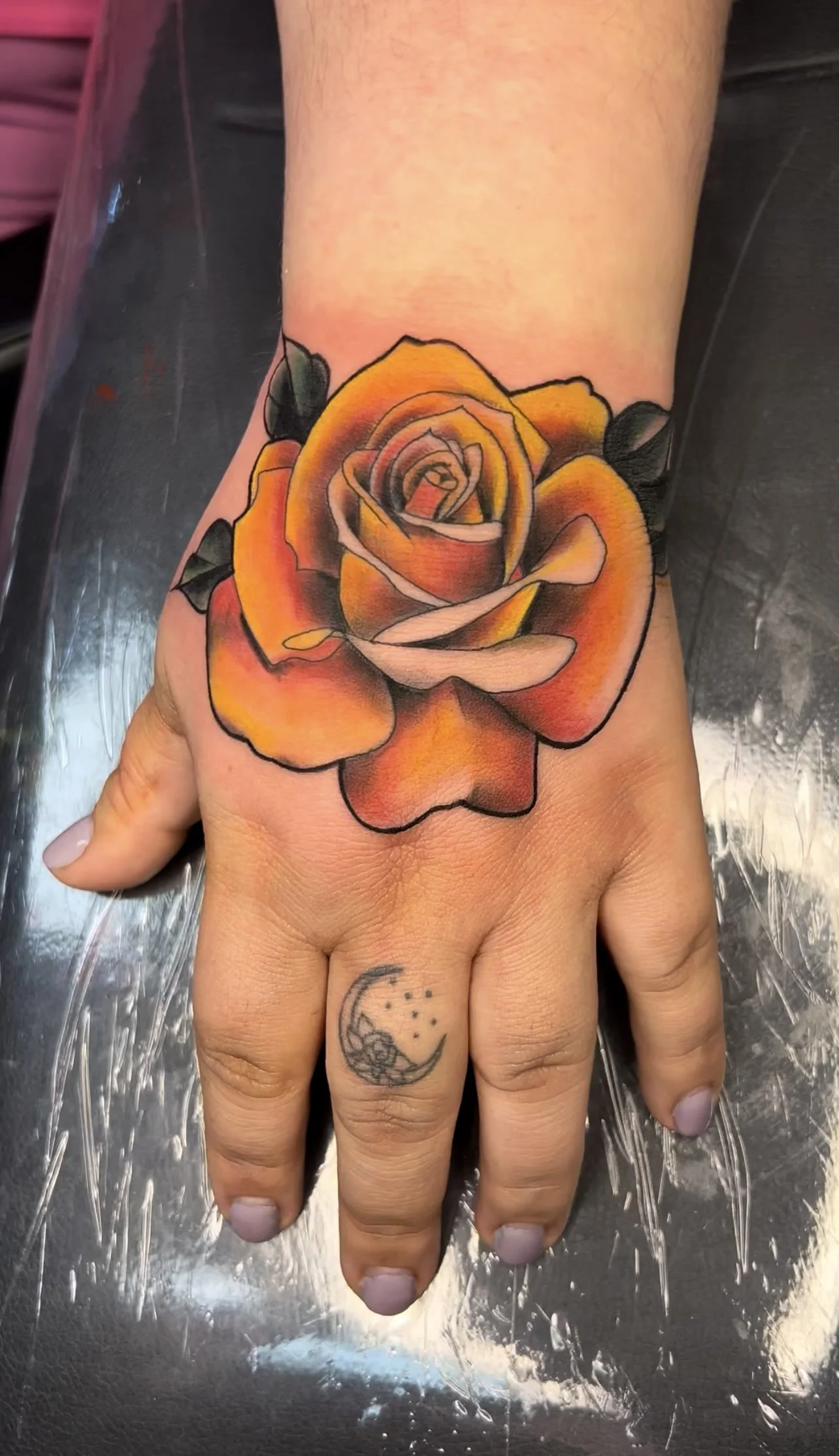 A hand with a vibrant orange and yellow rose tattoo on the back and a small moon and star tattoo on the finger.