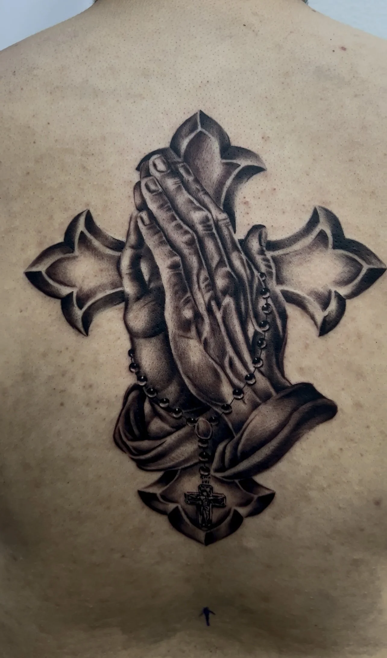 Black and gray tattoo of praying hands holding a rosary with a cross, superimposed over a Christian cross symbol, on a person's chest.