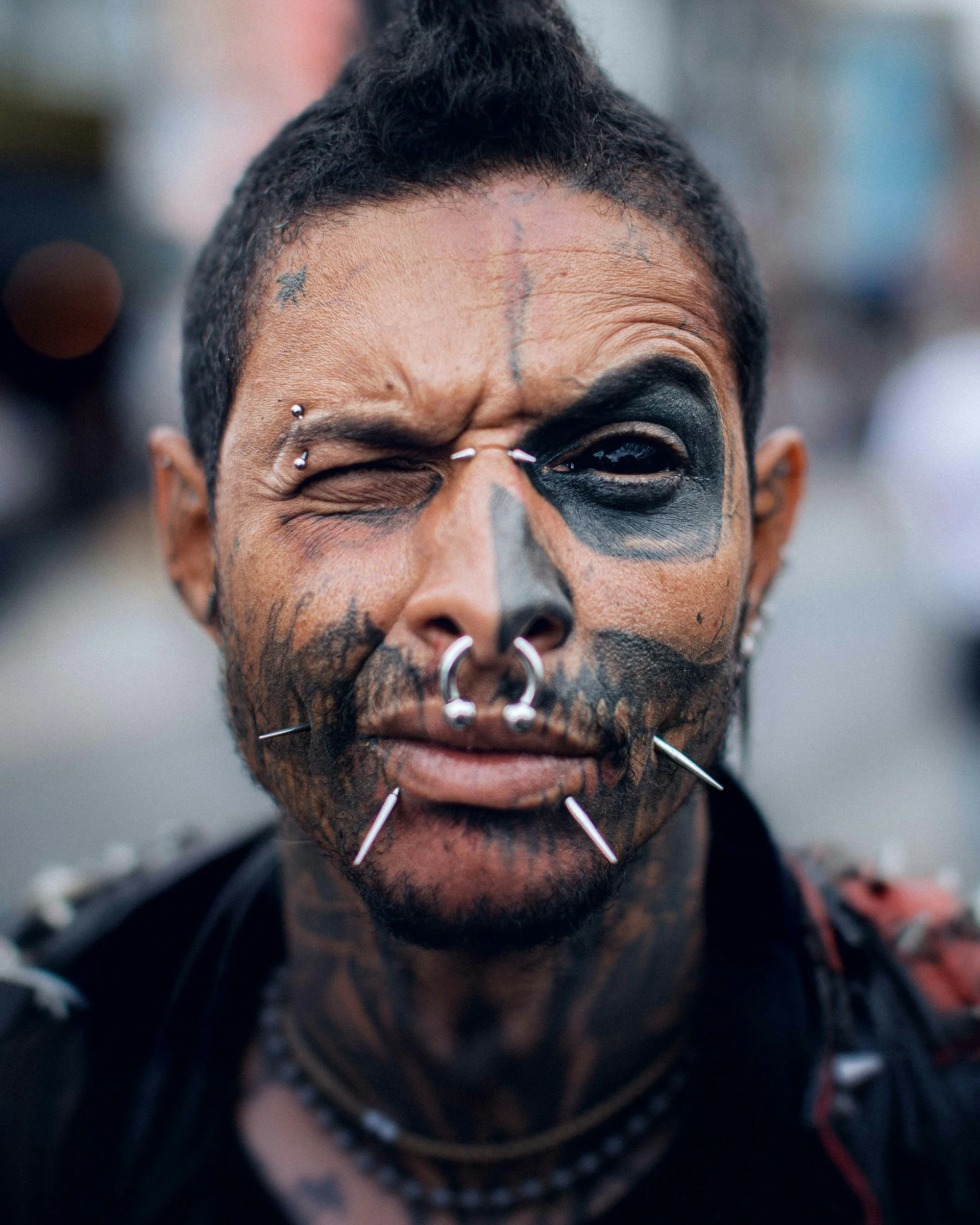 Close-up of a person's face with facial tattoos, piercings, and black makeup around one eye, winking with the other eye, wearing multiple nose and facial piercings and jewelry.