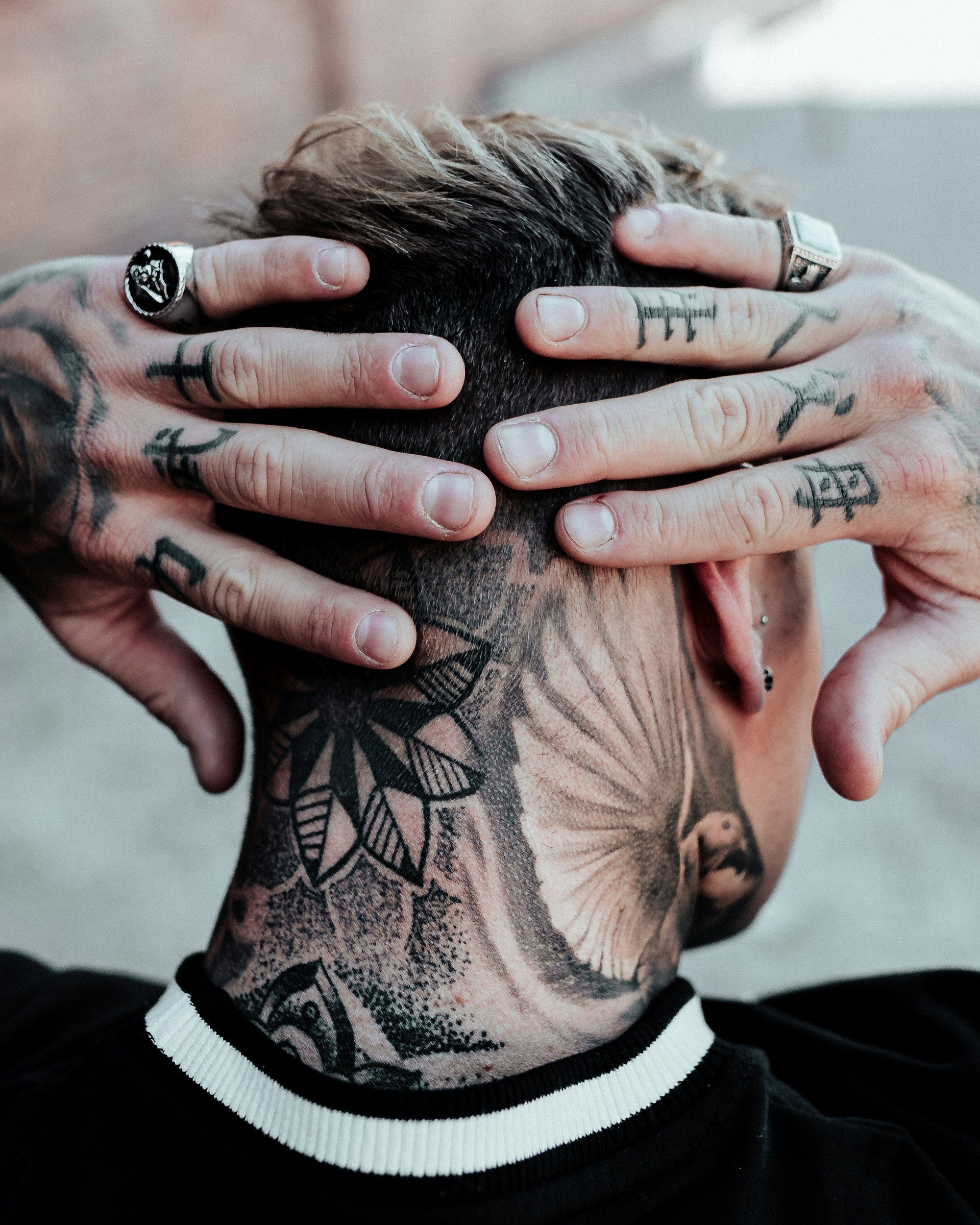 Close-up of a person's head being held with both hands. The person has short, dark hair with light-colored highlights. They are wearing multiple rings on their fingers, including one with a skull design. The person's hands are tattooed with various designs and lettering. The person is wearing a black and white striped shirt or sweater.