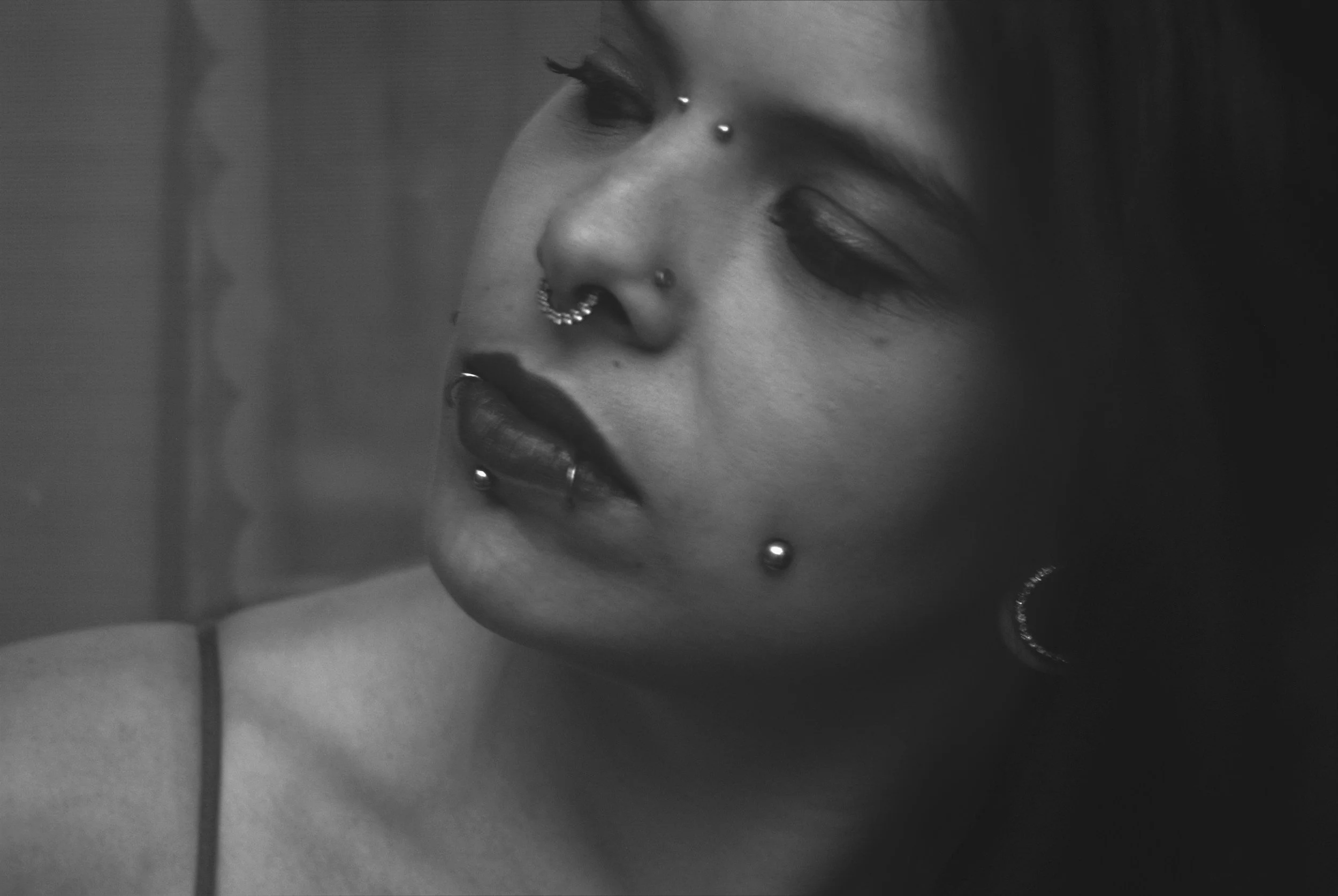 Black and white close-up of a woman's face with multiple facial piercings, including a septum ring, a nose stud, and earrings. She has dark makeup and long eyelashes.