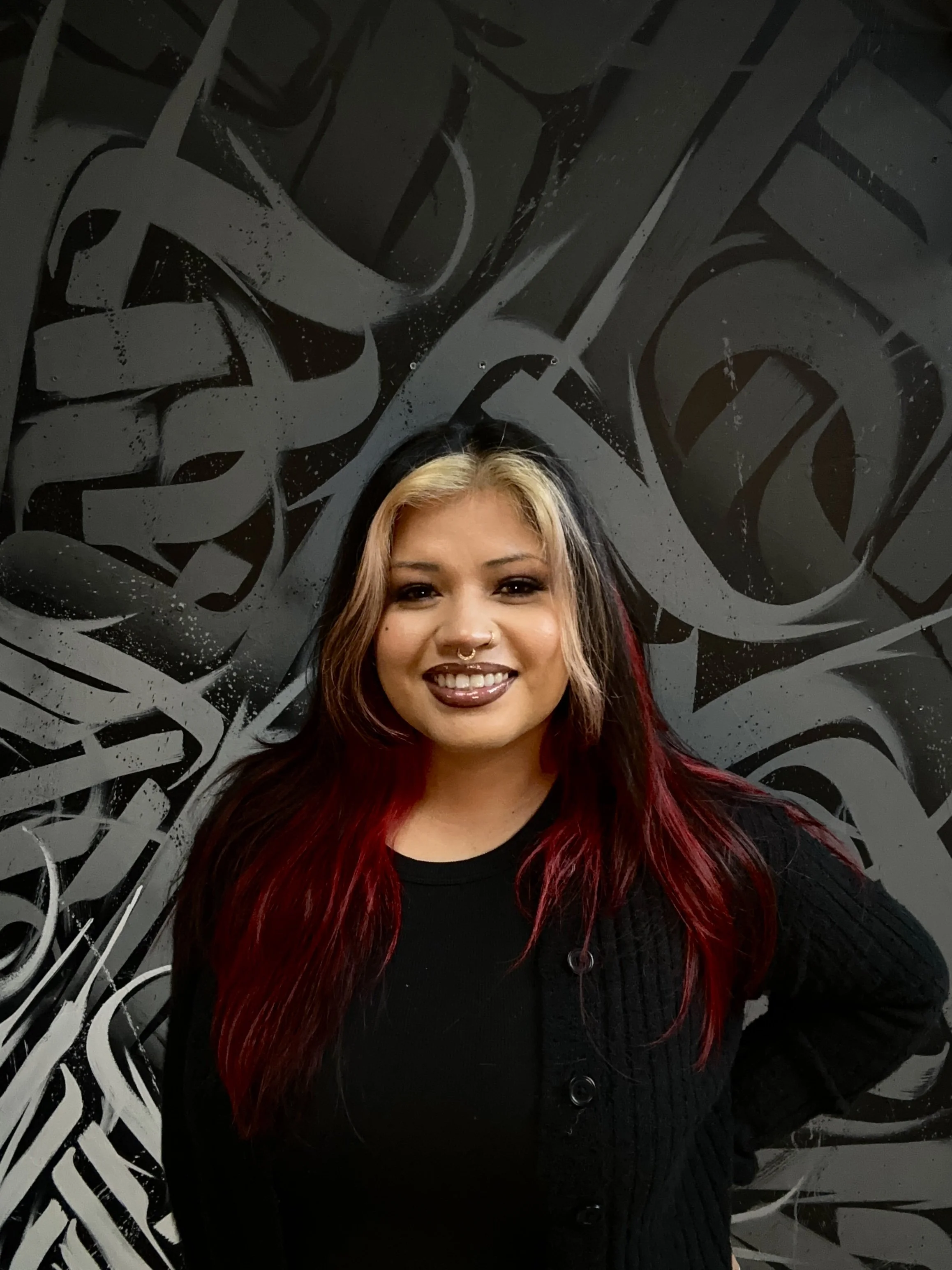 A woman with long hair that is blonde at the top and red at the bottom, smiling with dark lipstick, wearing a black top and black cardigan, standing in front of a black and white graffiti wall.