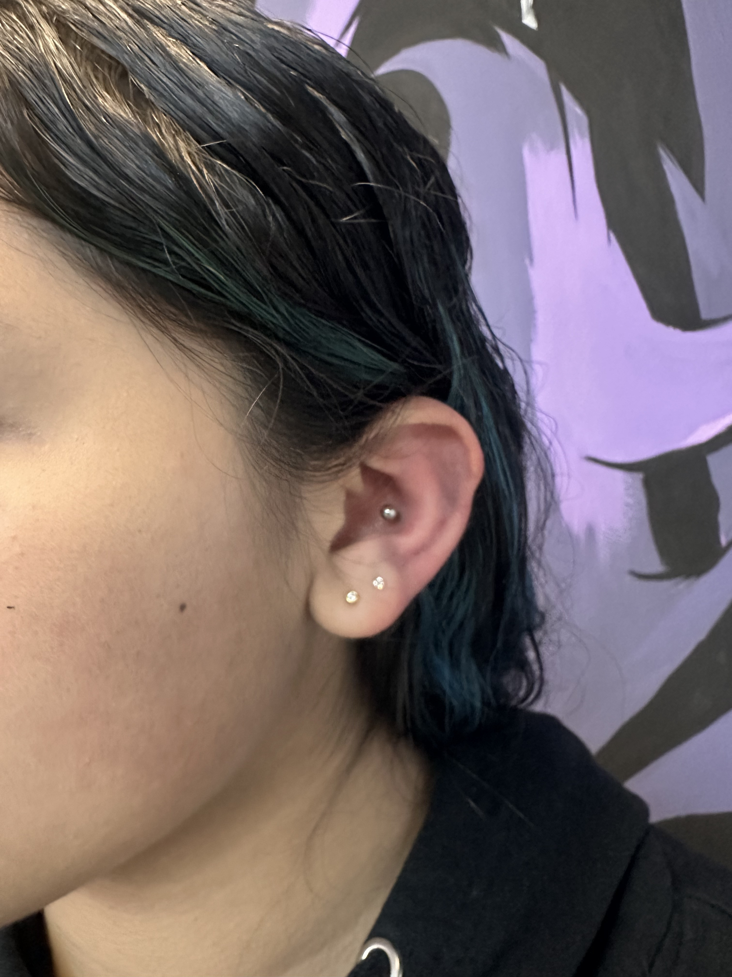 Close-up of a person's ear with multiple small earrings against a purple background with black abstract shapes.