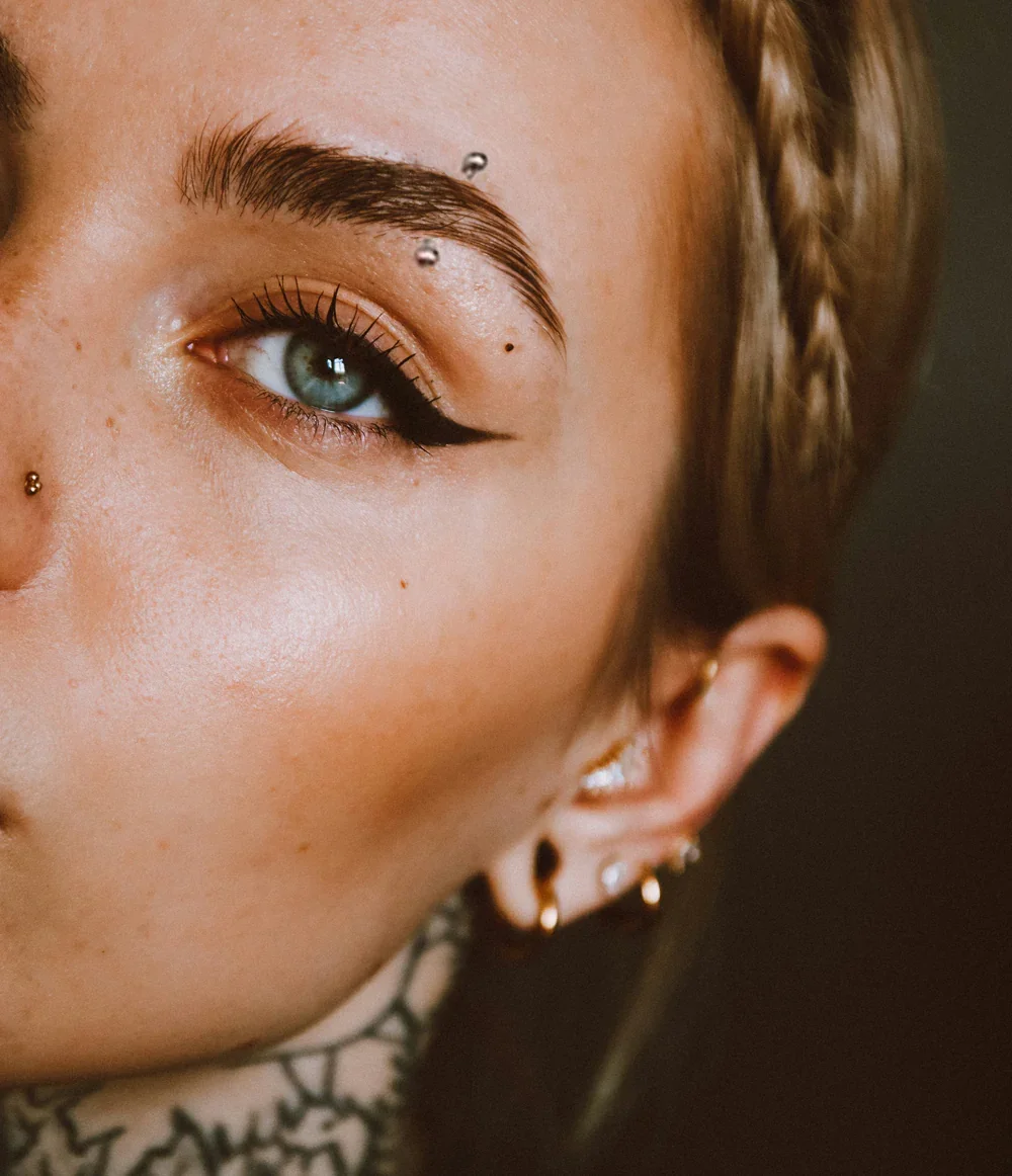 Close-up of a woman with blue eyes, dark eyeliner, multiple facial piercings, and earrings, with a small braid in her hair.
