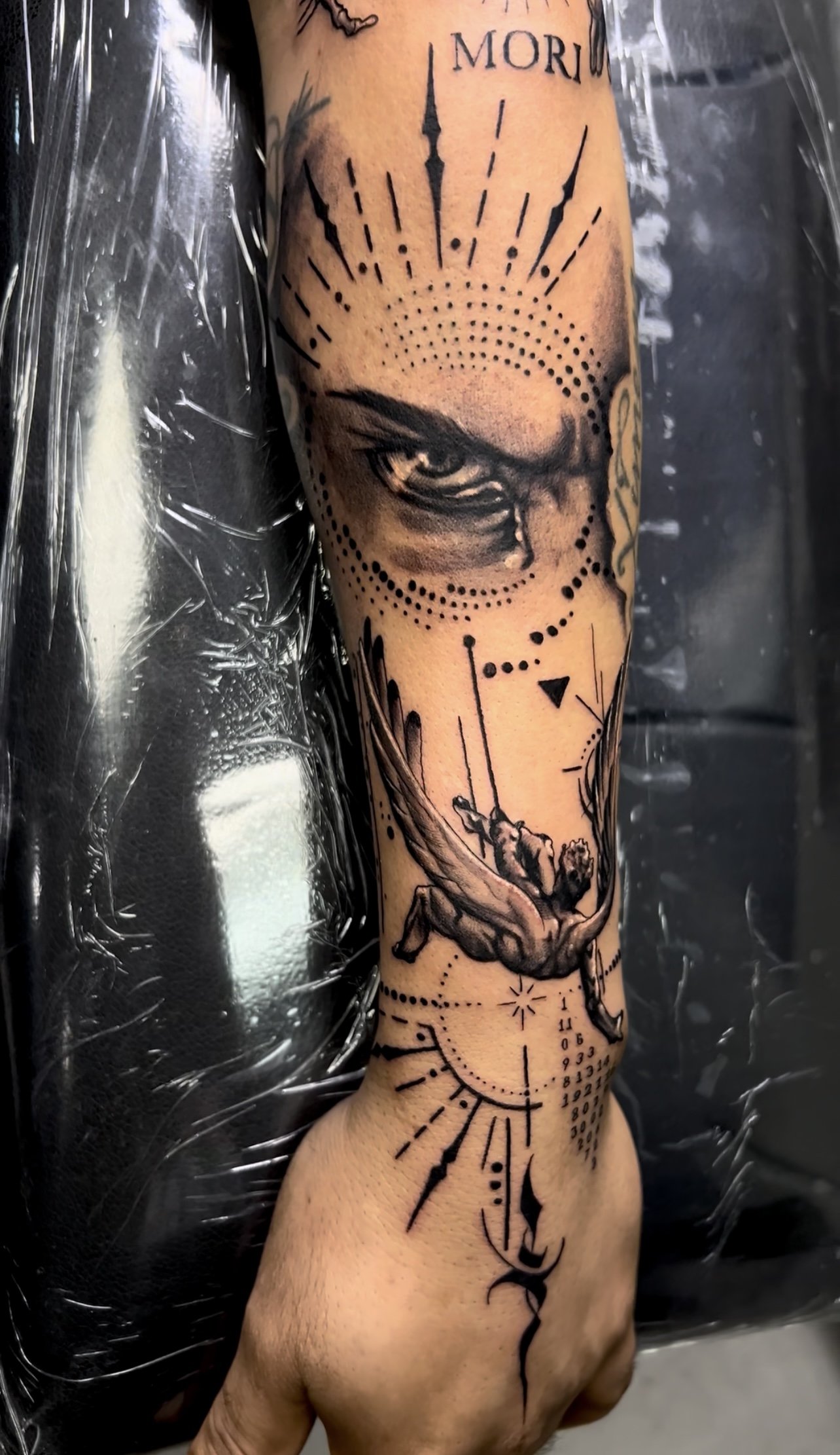 A tattoo on a person's forearm featuring an eye, a swinging figure, and various geometric and abstract elements.