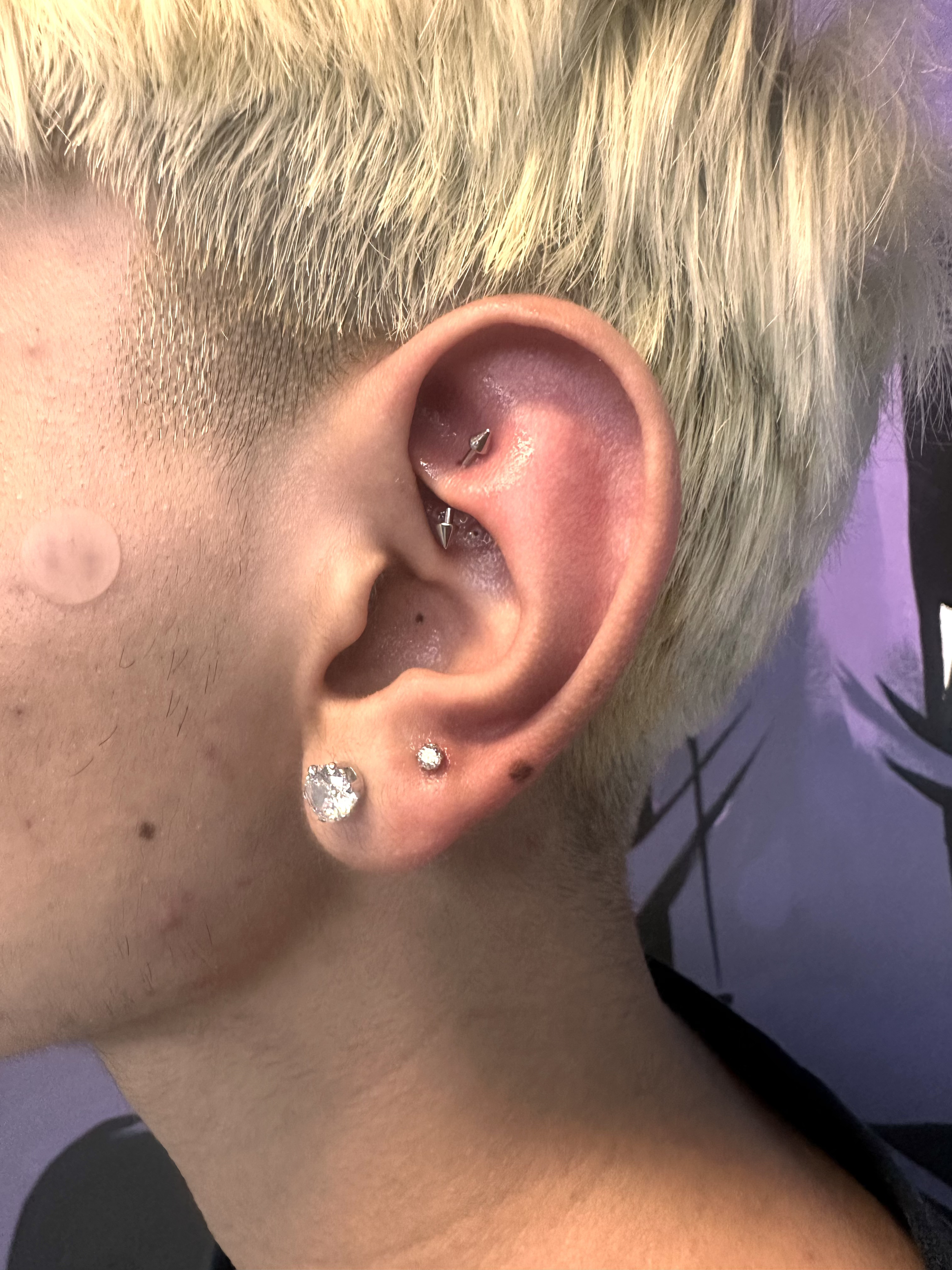 Close-up of a person's ear with multiple piercings, including a dangle earring, a small stud, a cartilage piercing with a hoop, and a small gemstone stud, with platinum blonde hair and visible skin freckles and moles.