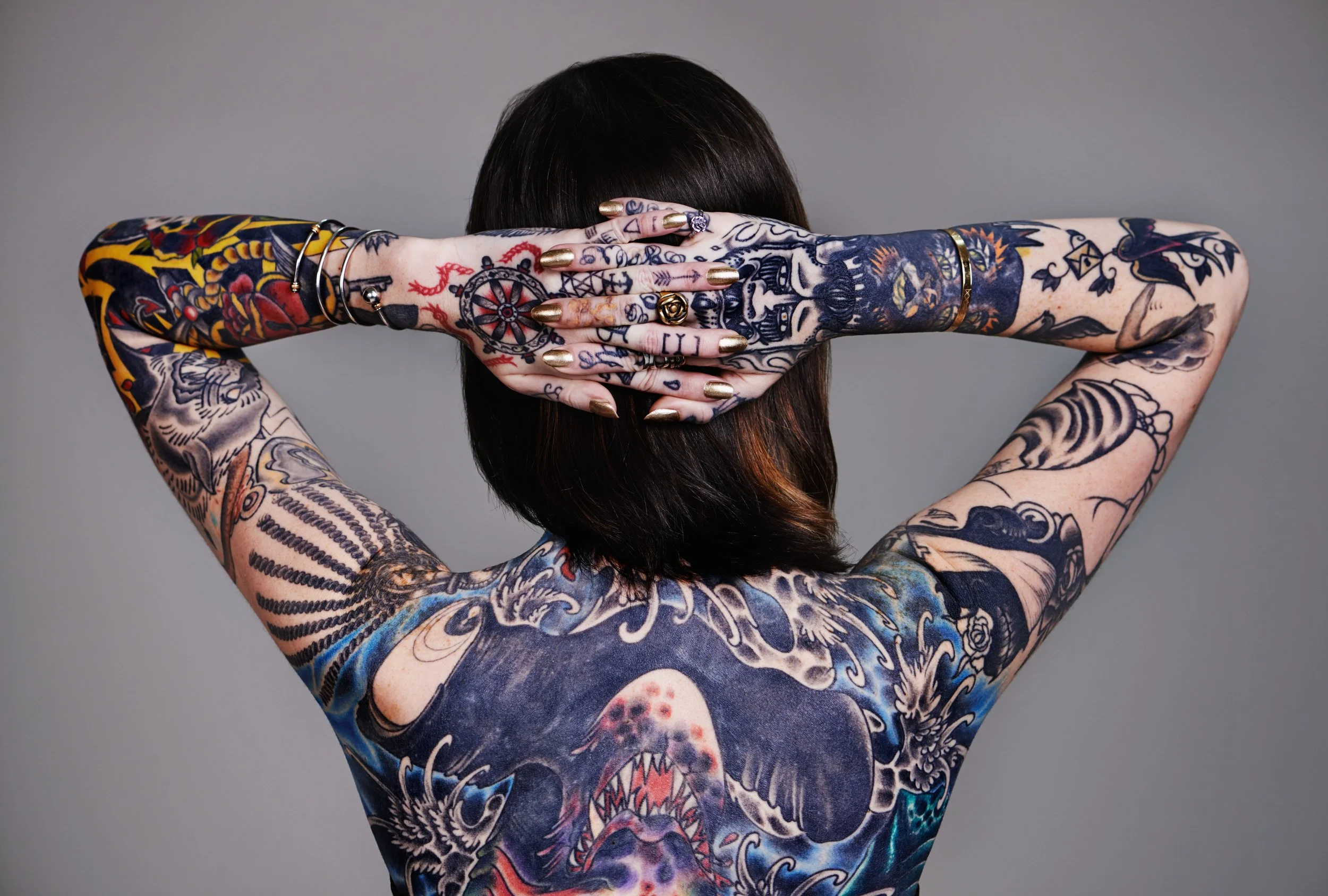 Back view of a tattooed woman with arms crossed behind her head, showing detailed tattoos on her arms, back, and shoulders, with short dark hair.