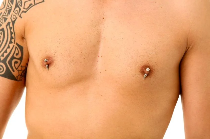 Close-up of a person's chest with nipple piercings and tribal tattoo on the left shoulder.