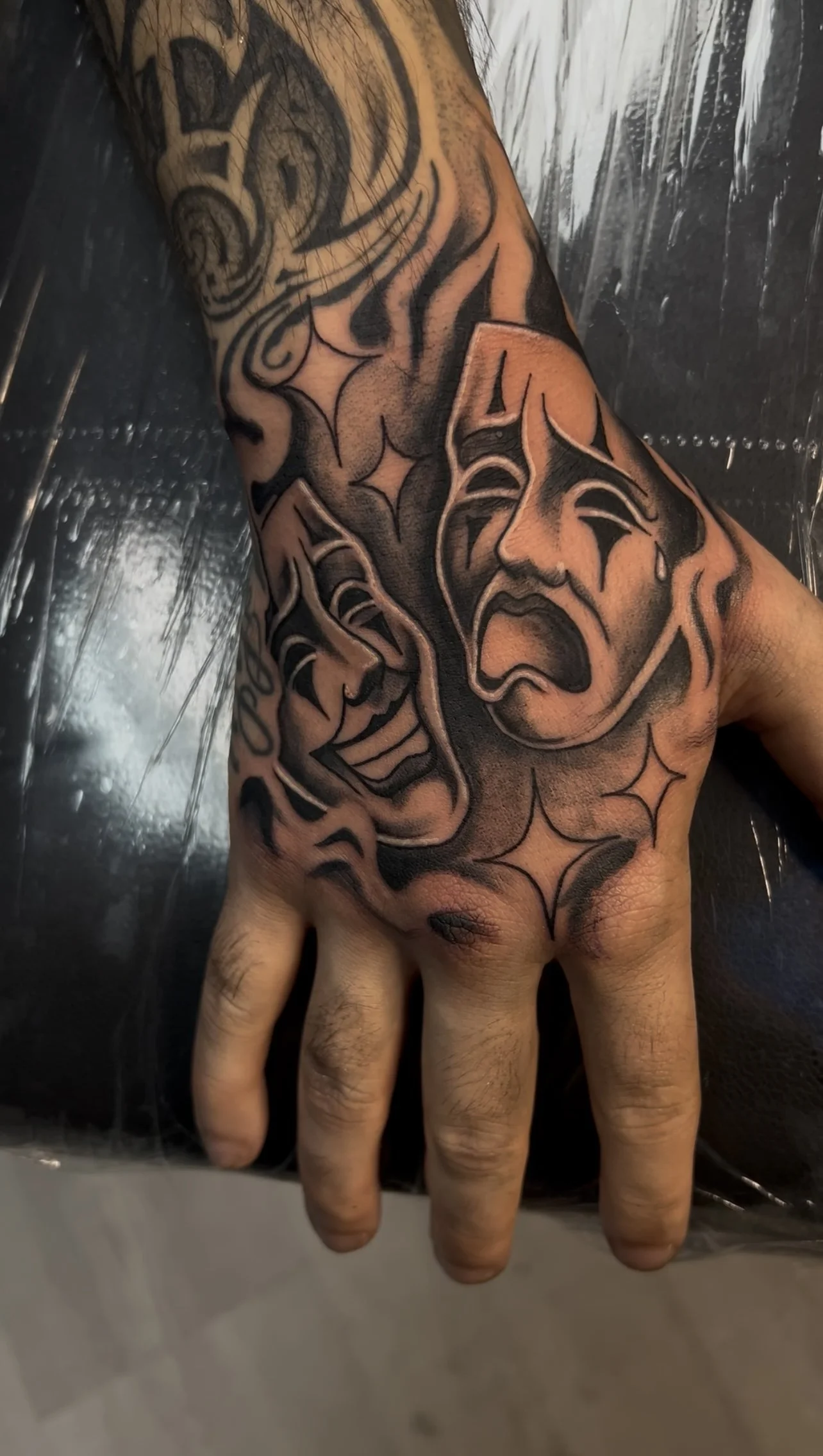Tattoo on hand featuring theatrical masks, one smiling and one crying, with stars and swirling designs.