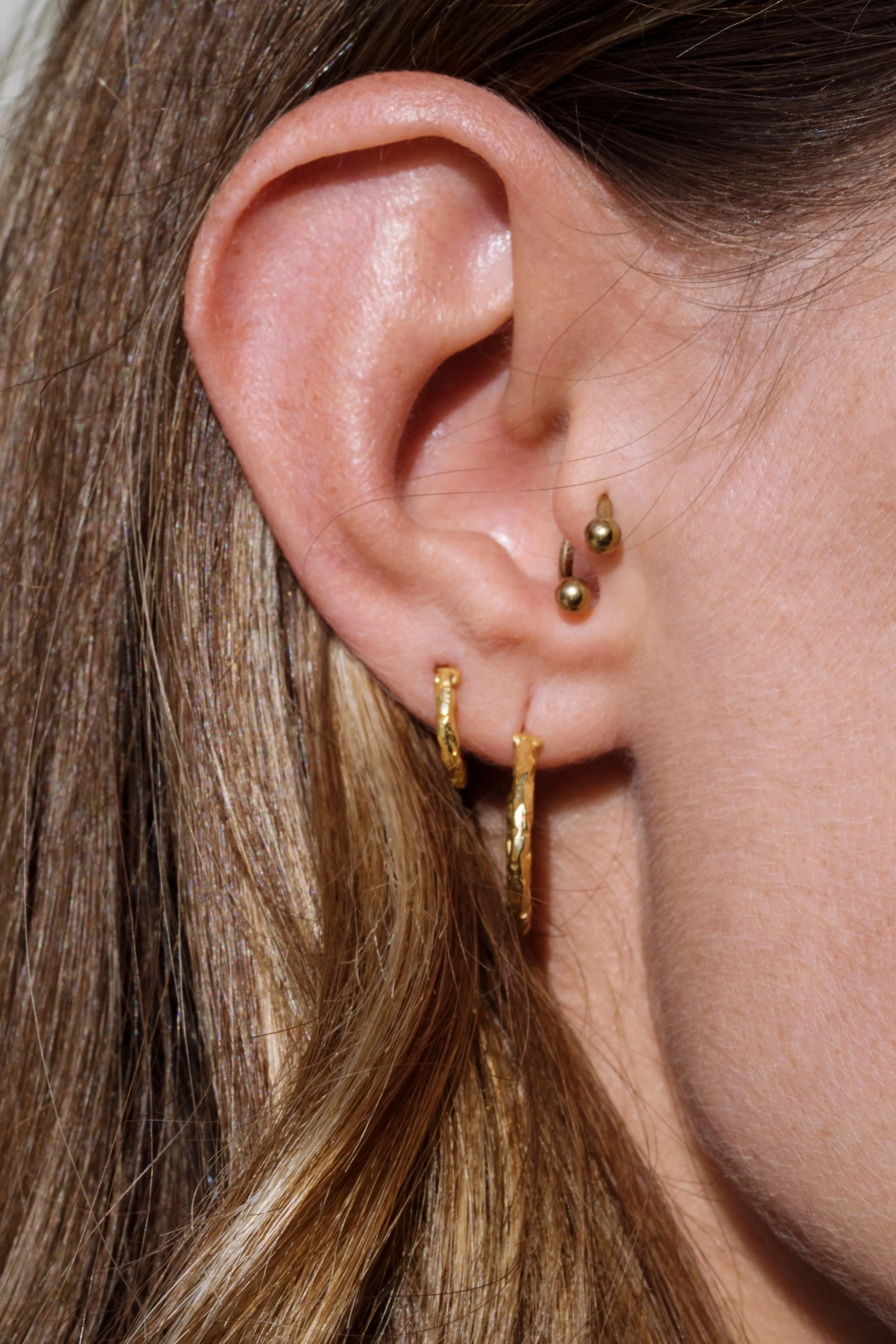 Close-up of a person's ear with multiple gold earrings, including a small hoop and a stud, and long brown hair.