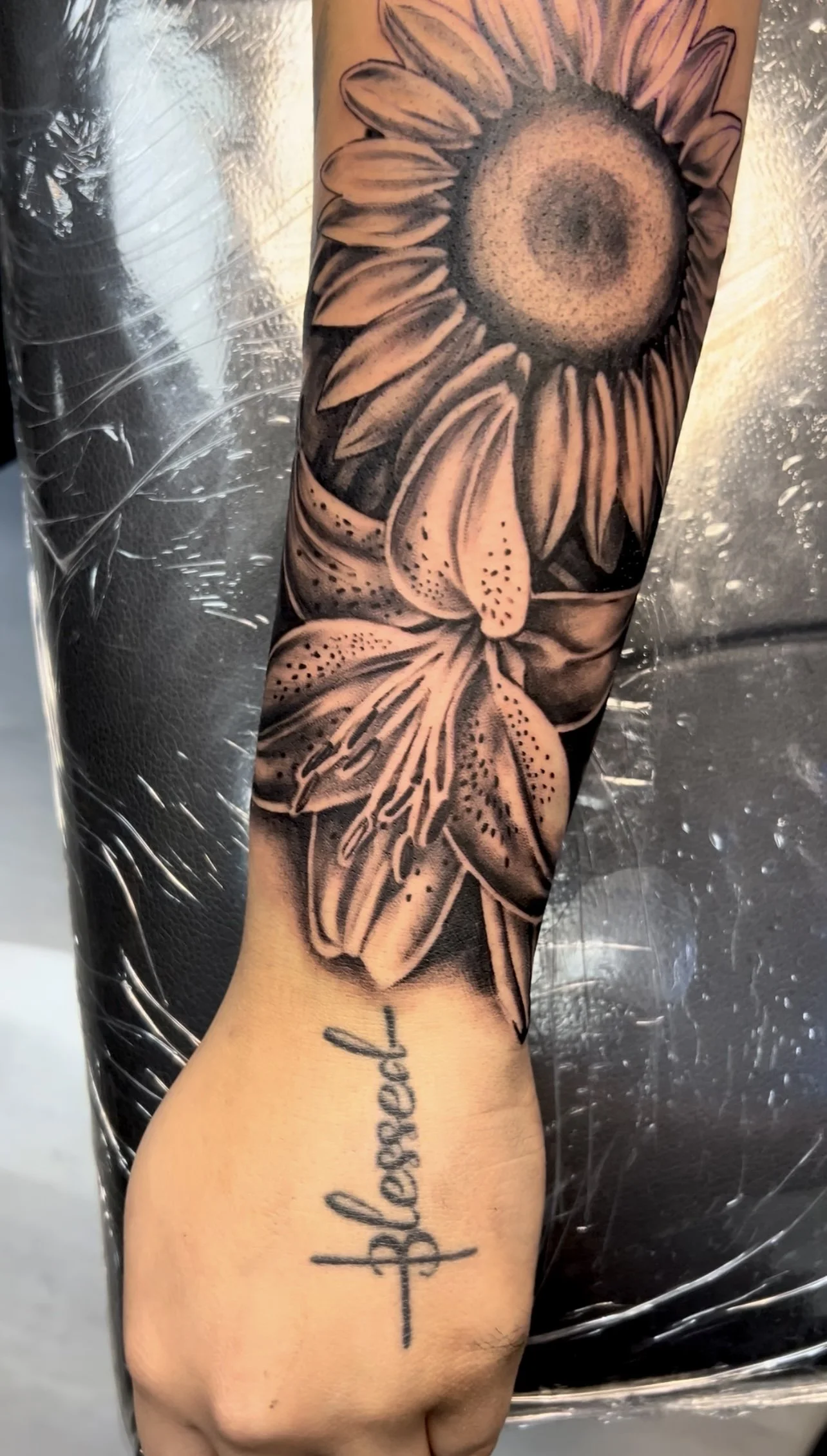 A black and gray tattoo of a sunflower and a lily on a person's forearm, with a handwritten-style word 'blessed' underneath.