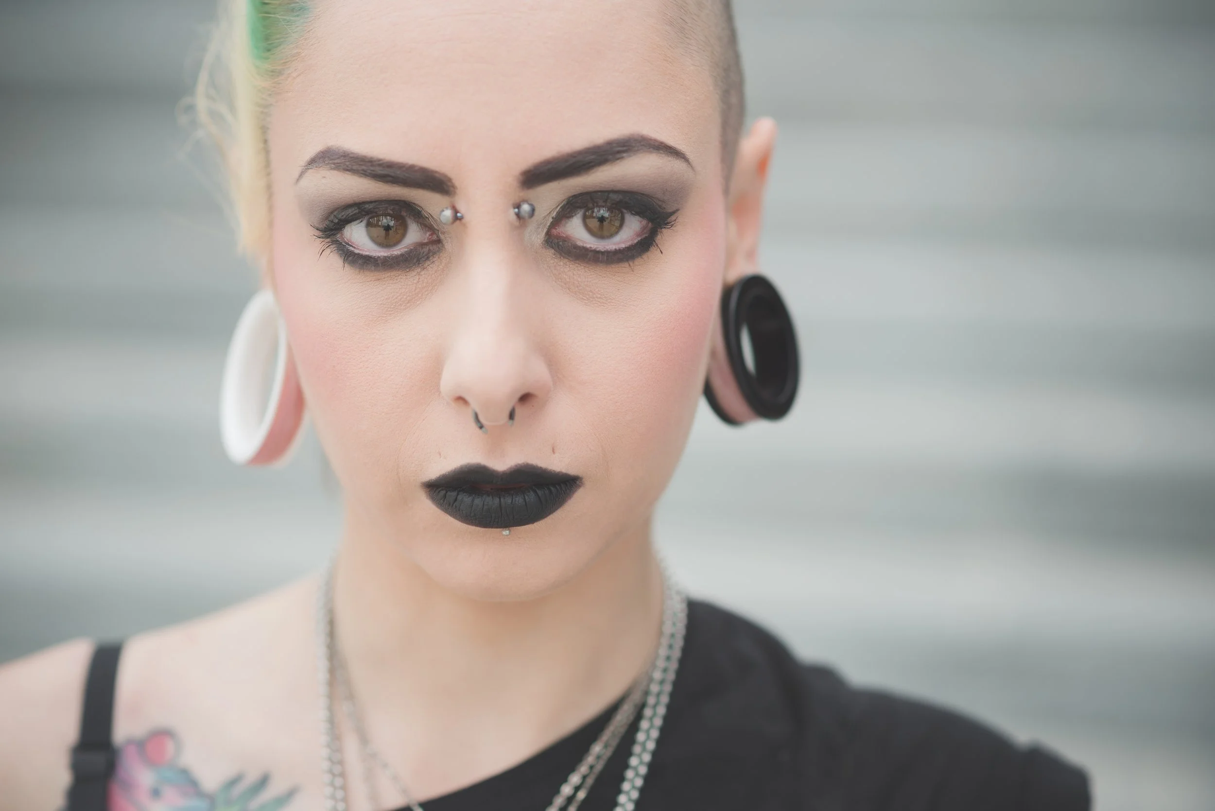 A woman with bold makeup and multiple facial piercings. She has dark lipstick, eye makeup, and facial jewelry, including a septum ring and eyebrow studs. She is wearing a black top with straps, a pearl necklace, and has colorful tattoos on her shoulder.