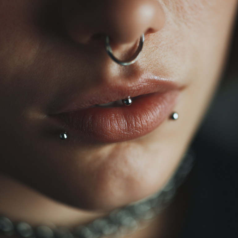 Close-up of a person's lips with a septum ring and two lip piercings, one on the lower lip and one on the side, with edited dark background.