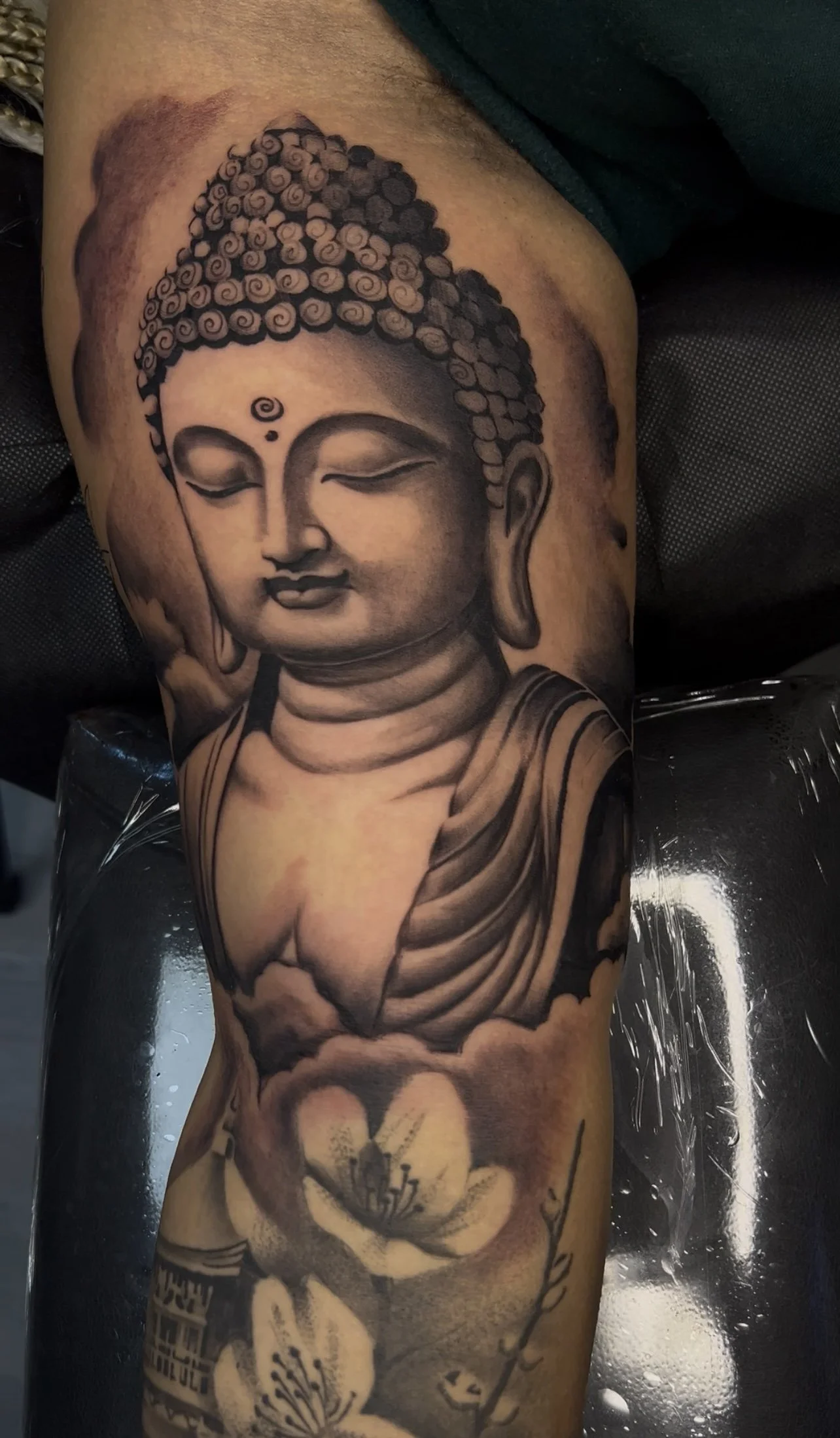 Black and gray tattoo of Buddha's face with closed eyes, curly hair, and a small spiral on the forehead, with lotus flowers below.