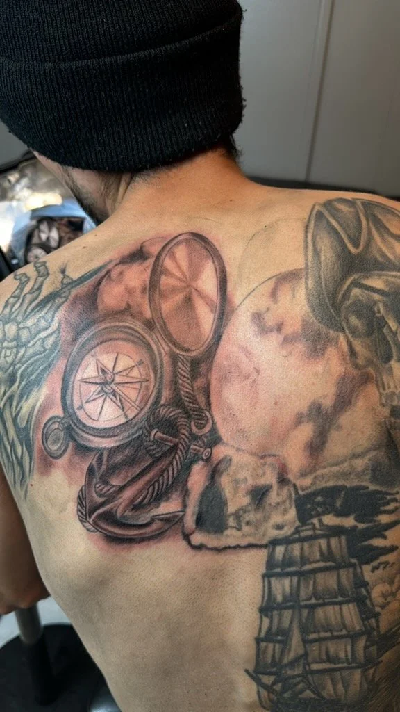Tattoo on person's shoulder and chest featuring a compass, anchor, moon, skull, and sailing ship in black and gray ink.