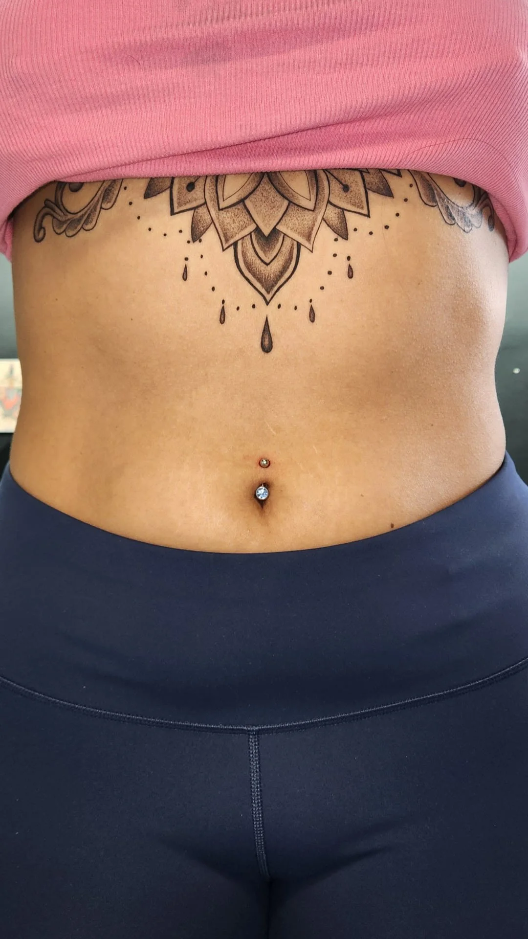 Close-up of a person's midsection showing a large tattoo of a floral design and hanging droplets on the upper abdomen, a belly button piercing with two jewels, a pink crop top, and dark athletic leggings.