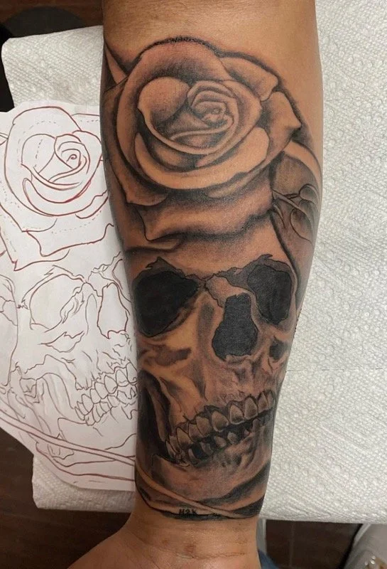 Black and gray tattoo of a skull with a rose on top, on a person's forearm.