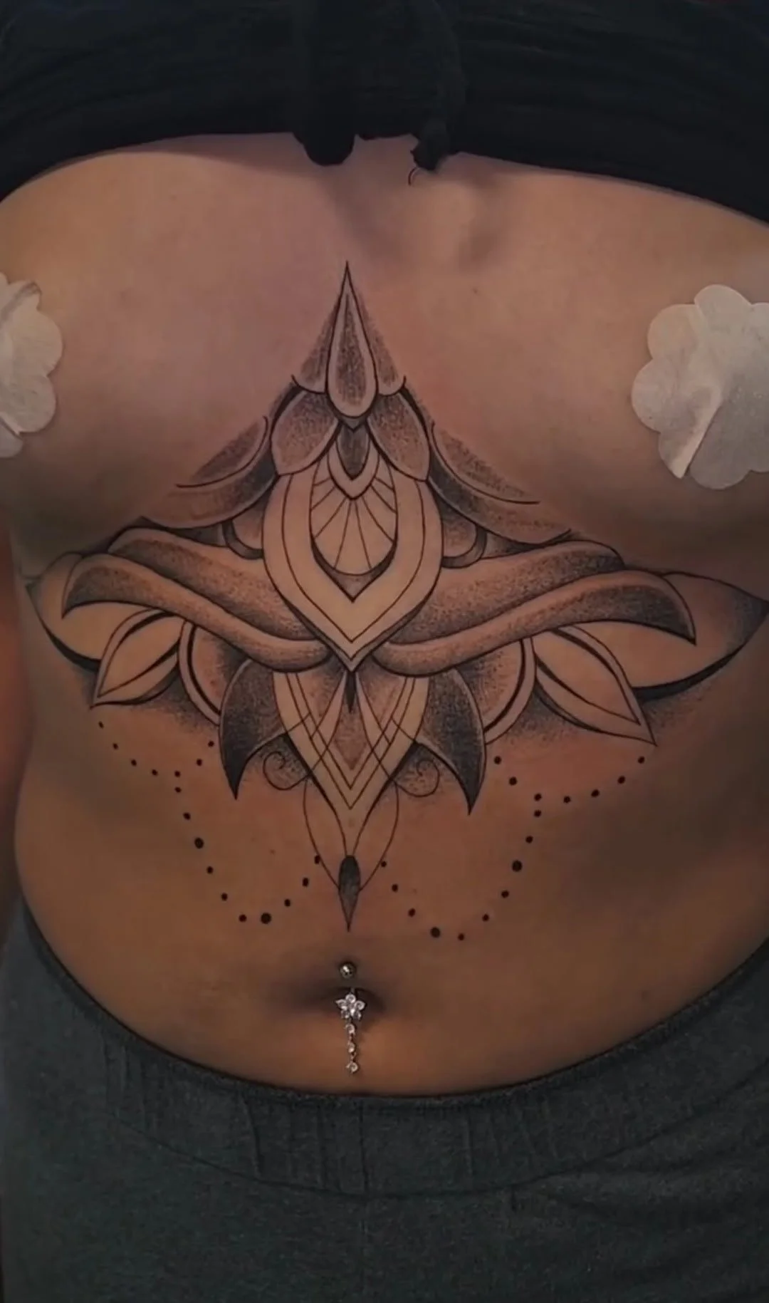 Close-up of a tattoo on a person's chest featuring a symmetrical, intricate, geometric floral design with dots and shading, and white 3D flower embellishments on the shoulders.