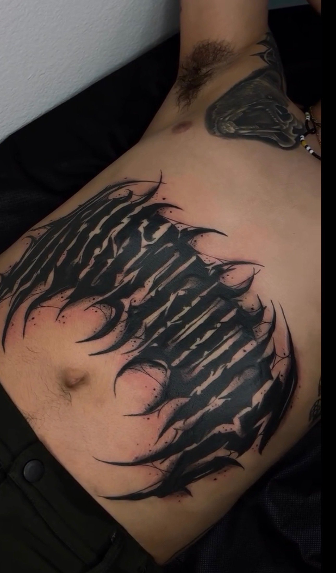 A close-up of a person's torso and part of their arm, showcasing a large, black, abstract tattoo design on the chest and side, with additional tattoos visible on the arm and neck.