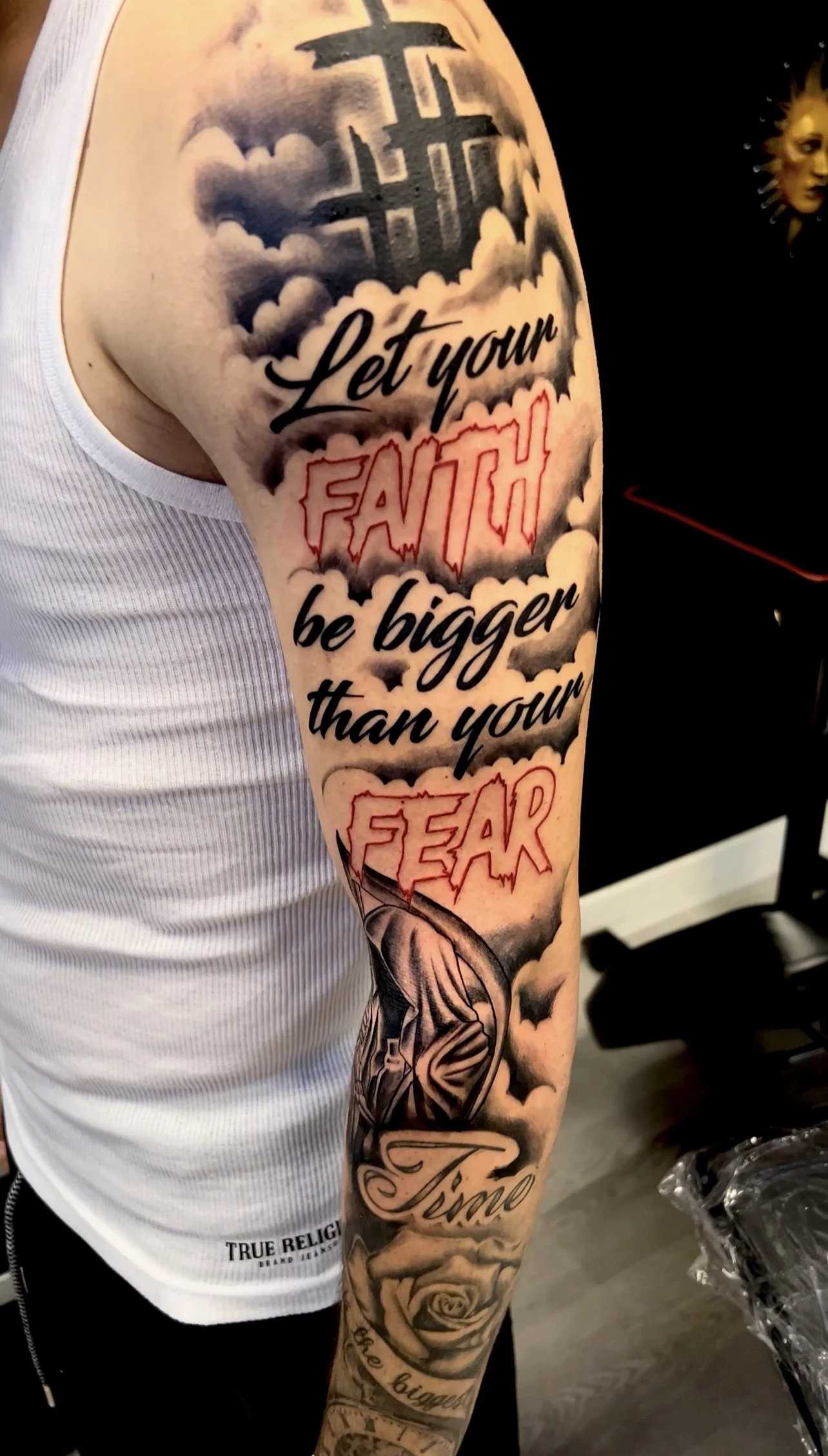 Tattoo on a person's arm with clouds and sky in the background, featuring the phrase 'Let your FAITH be bigger than your FEAR' in stylized fonts and additional tattoo designs at the bottom.