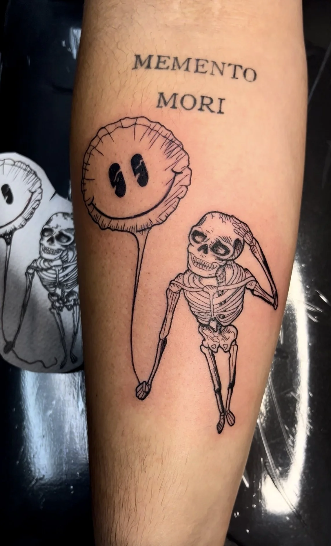 Tattoo of a smiling flower with eyes, a skeleton, and the phrase 'Memento Mori' above.