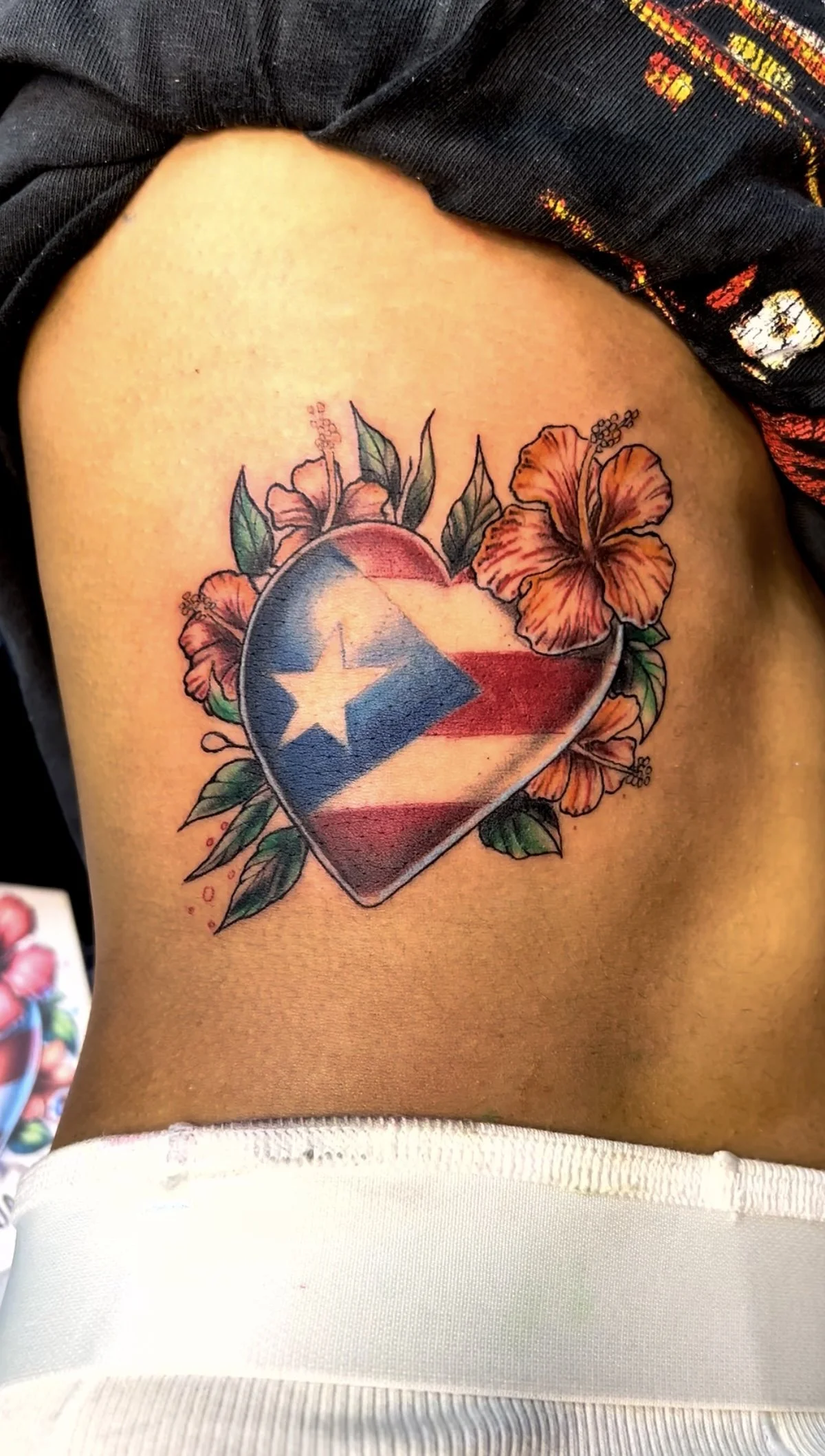 Tattoo of a heart with the Puerto Rican flag on it, surrounded by orange hibiscus flowers and green leaves.
