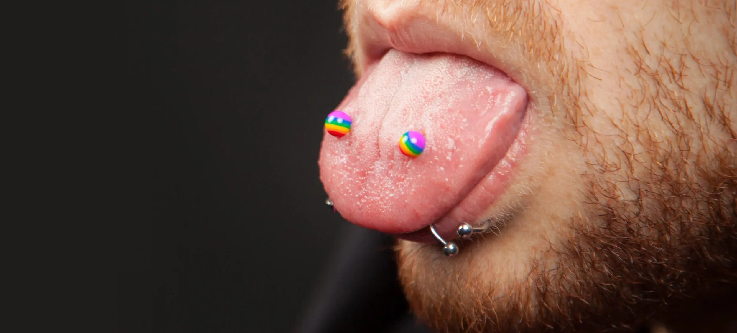 Close-up of a person's tongue with rainbow-colored ball-shaped tongue piercing, additional pierced jewelry including a septum ring, with facial hair and a dark background.