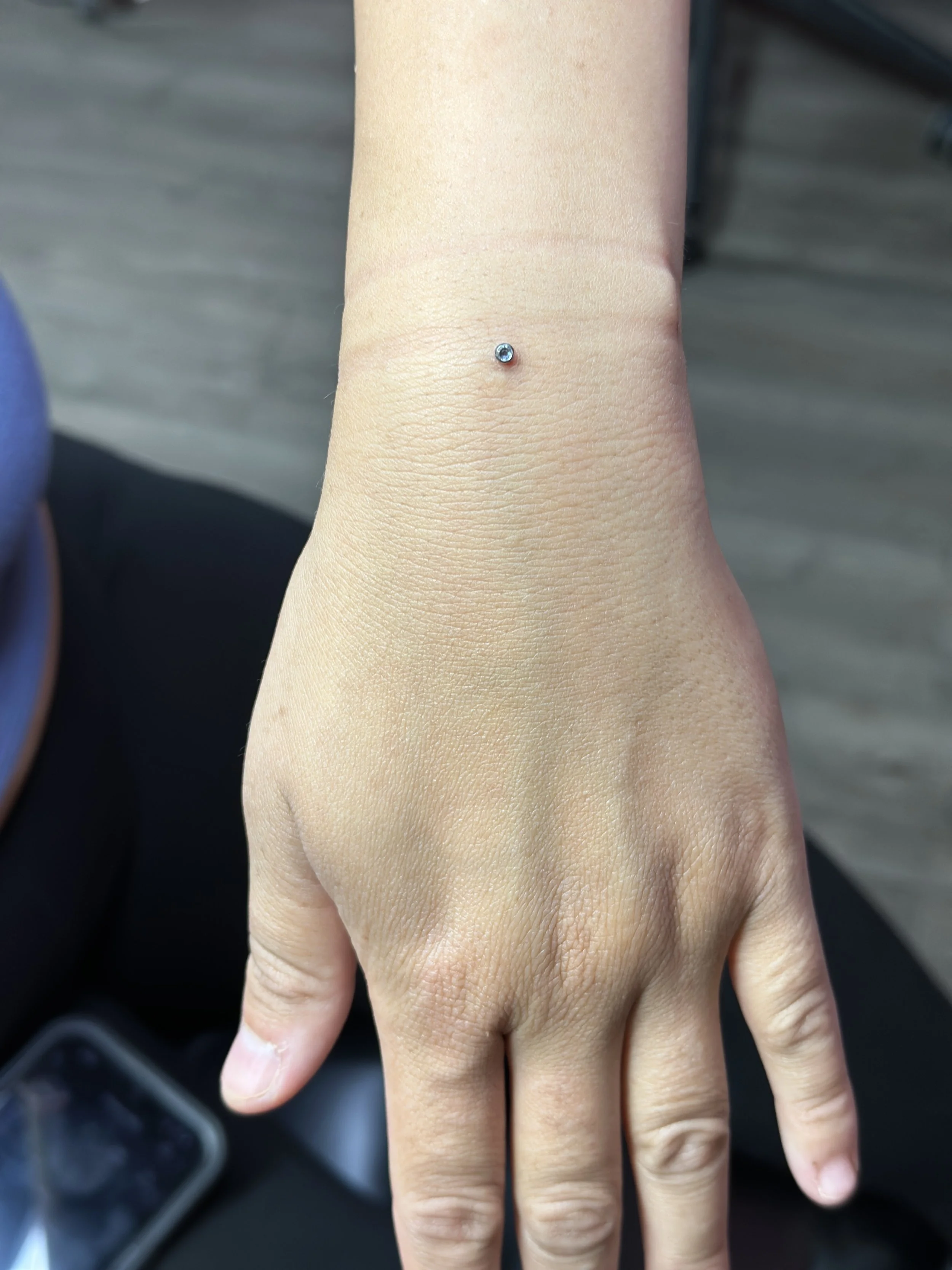 A close-up of a person's left hand and wrist, showing a small silver stud with a blue gemstone piercing on the wrist.