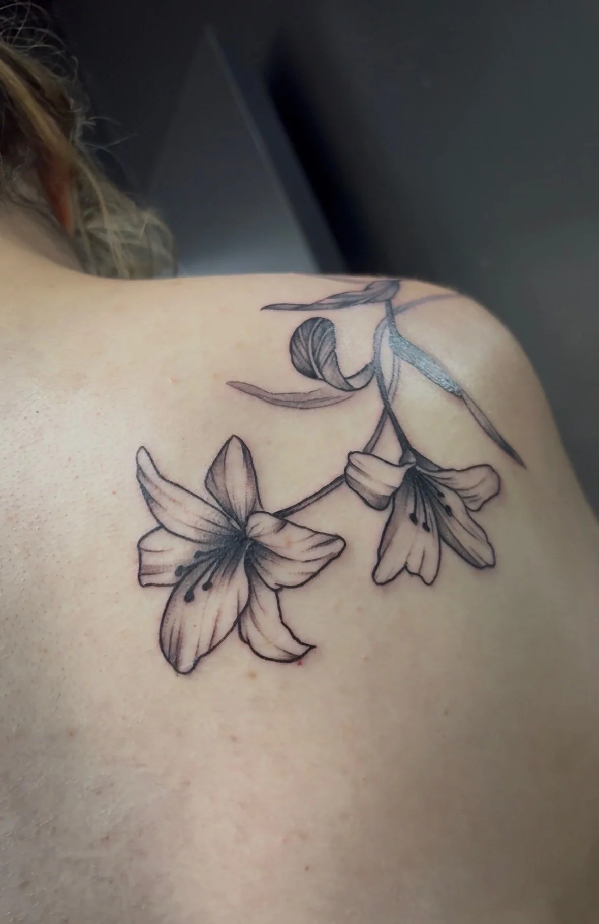 Black and gray tattoo of a branch with two lilies on a person's shoulder.