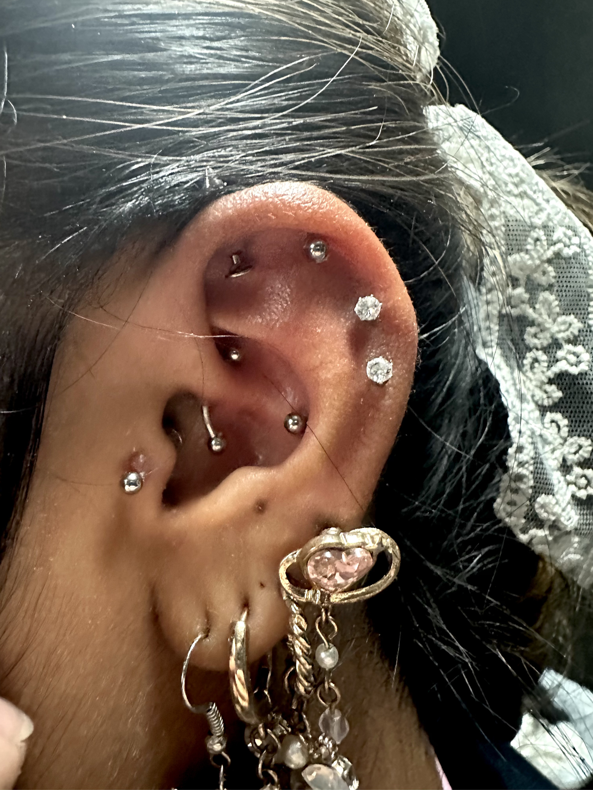 Close-up of a person's ear with multiple piercings and jewelry, including small stud earrings, a curved barbell, and a large decorative earring with a pink gemstone and dangling charms.