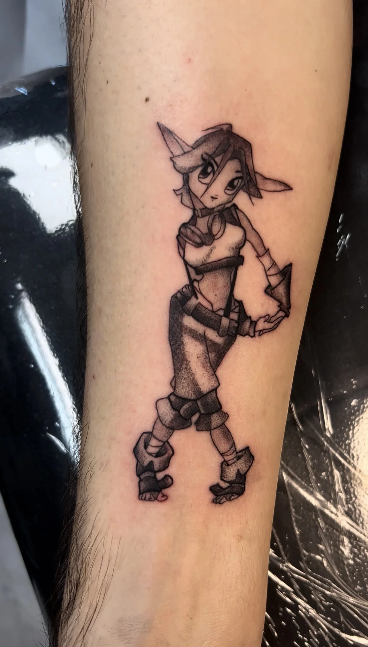 Black and gray tattoo of a female anthropomorphic fox character with large eyes, pointed ears, short hair, wearing a jacket, shorts, and boots, standing with hands on hips.