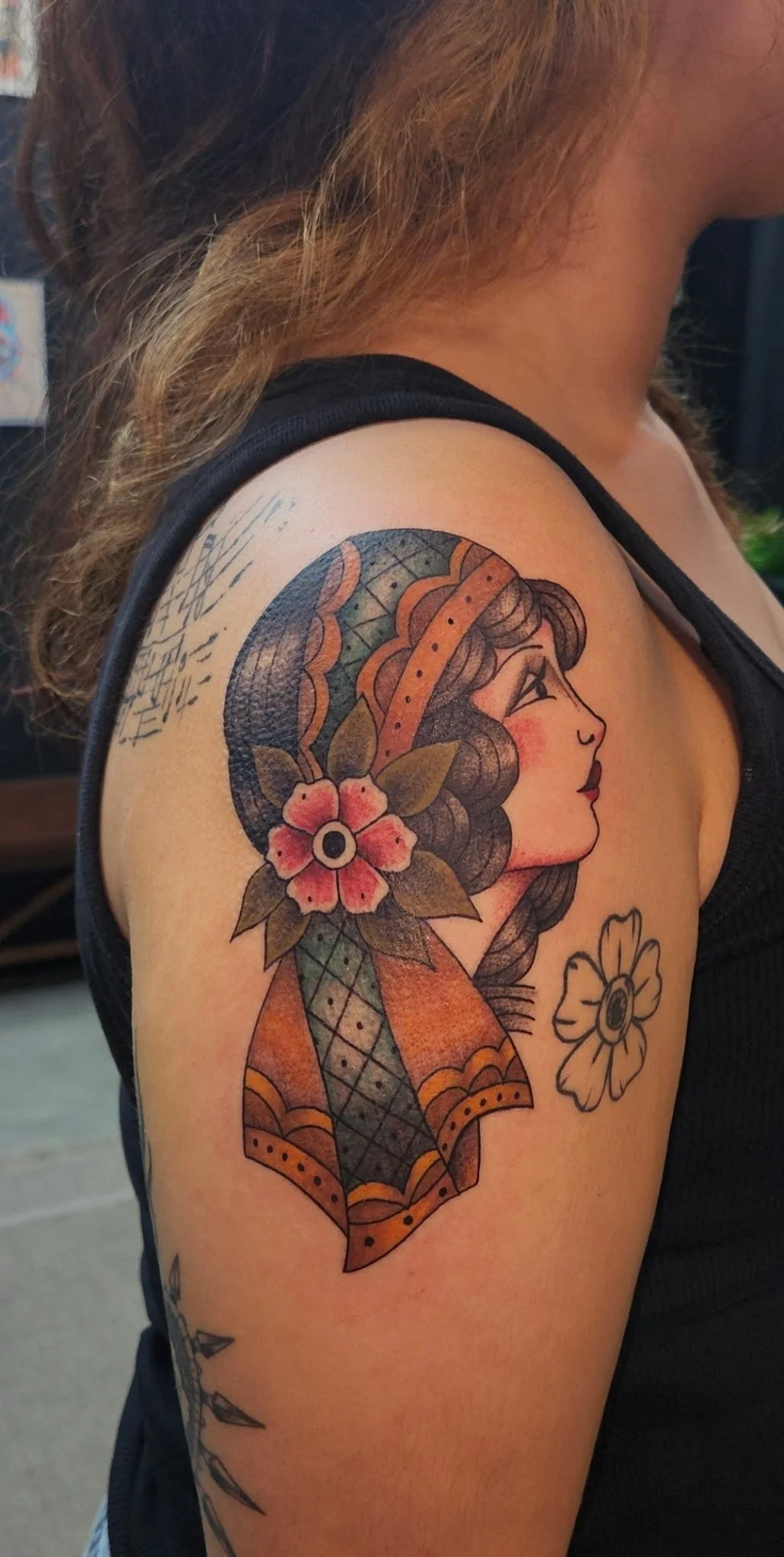 Tattoo of a woman's profile with long wavy hair, wearing a patterned headscarf, with a flower near her shoulder and a smaller flower below.