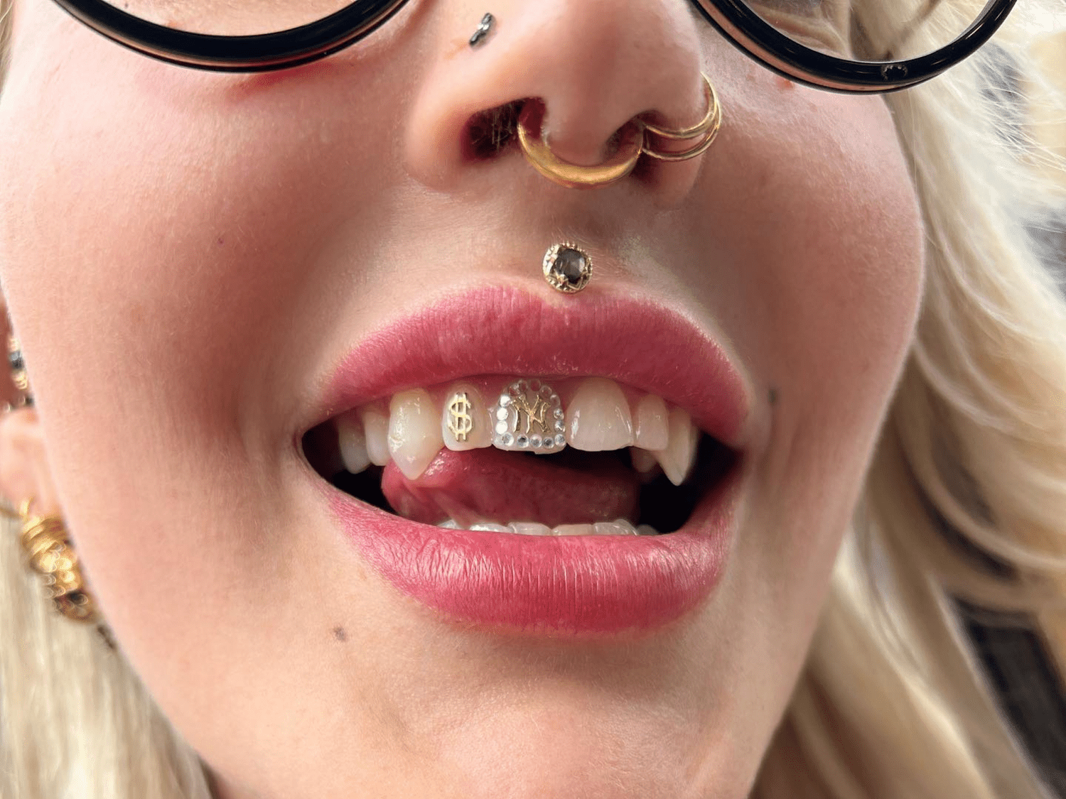 Close-up of a woman with blonde hair, wearing glasses, with multiple facial piercings, and colorful, jewelry-adorned teeth visible as she smiles.