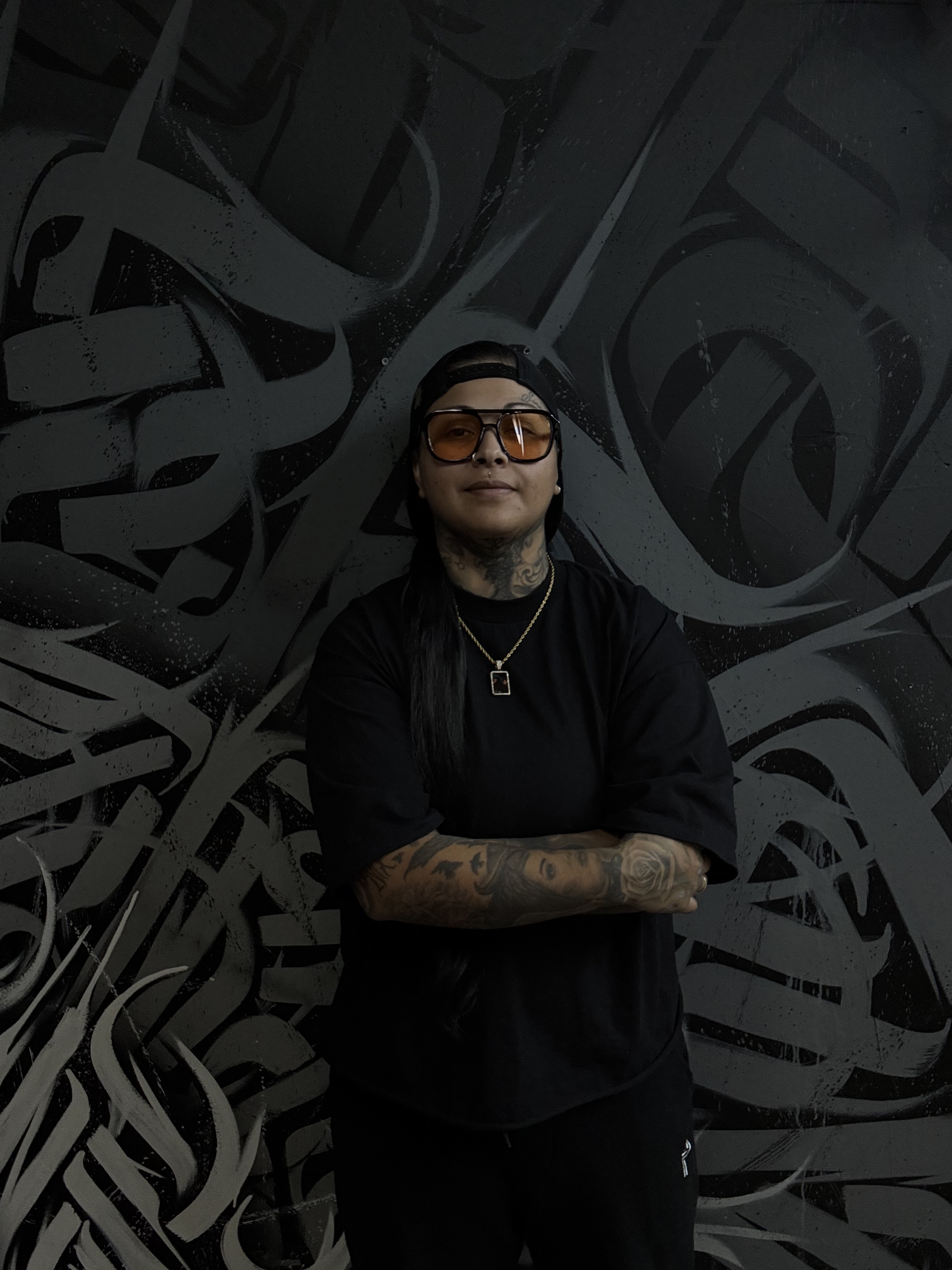 Person with long dark hair, tattoos on arms and neck, wearing sunglasses, a black cap backwards, black t-shirt, gold necklace with pendant, standing with arms crossed in front of a black and gray graffiti wall.