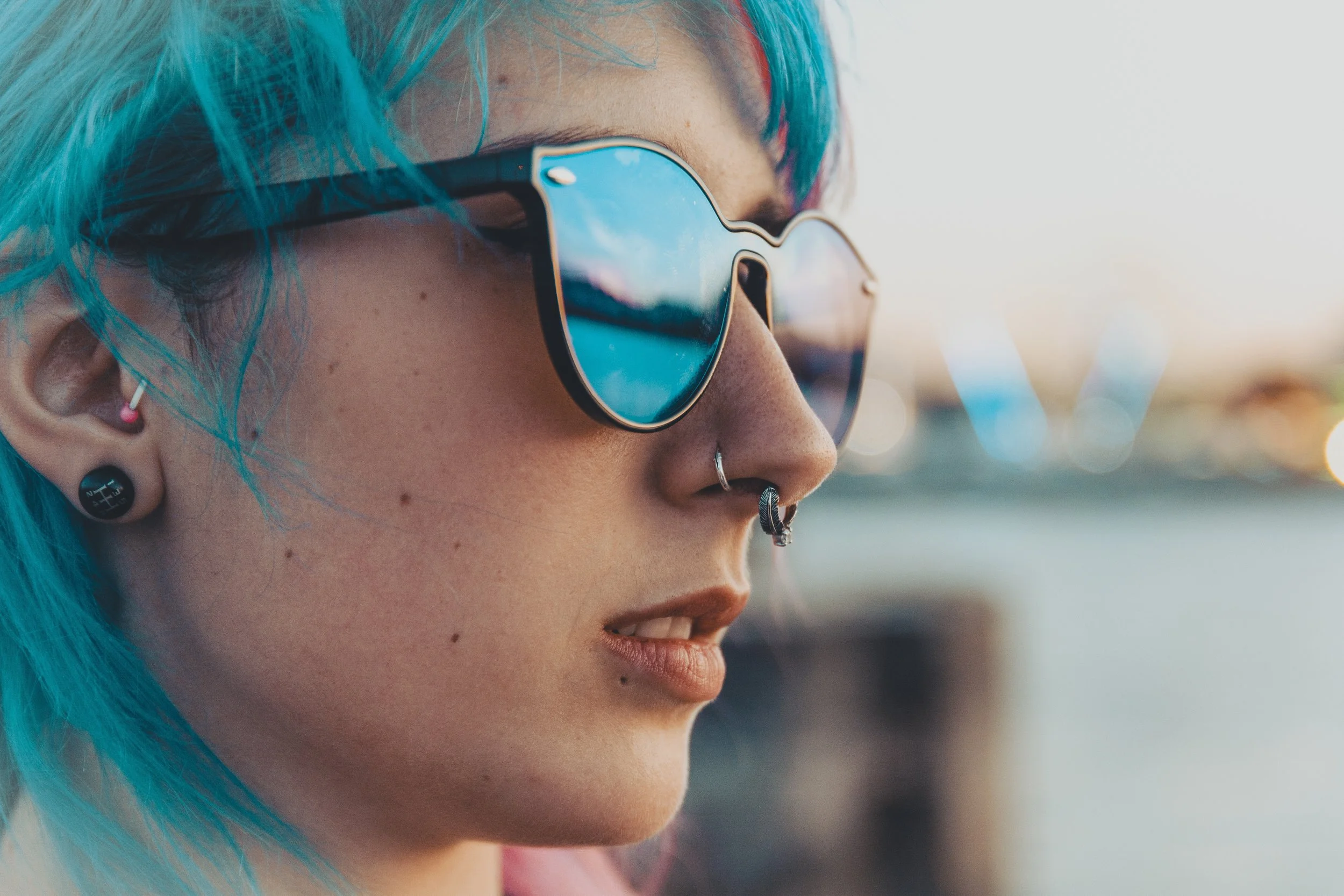 Close-up of a woman with bright blue hair wearing sunglasses and multiple earrings, with a blurred outdoor background.
